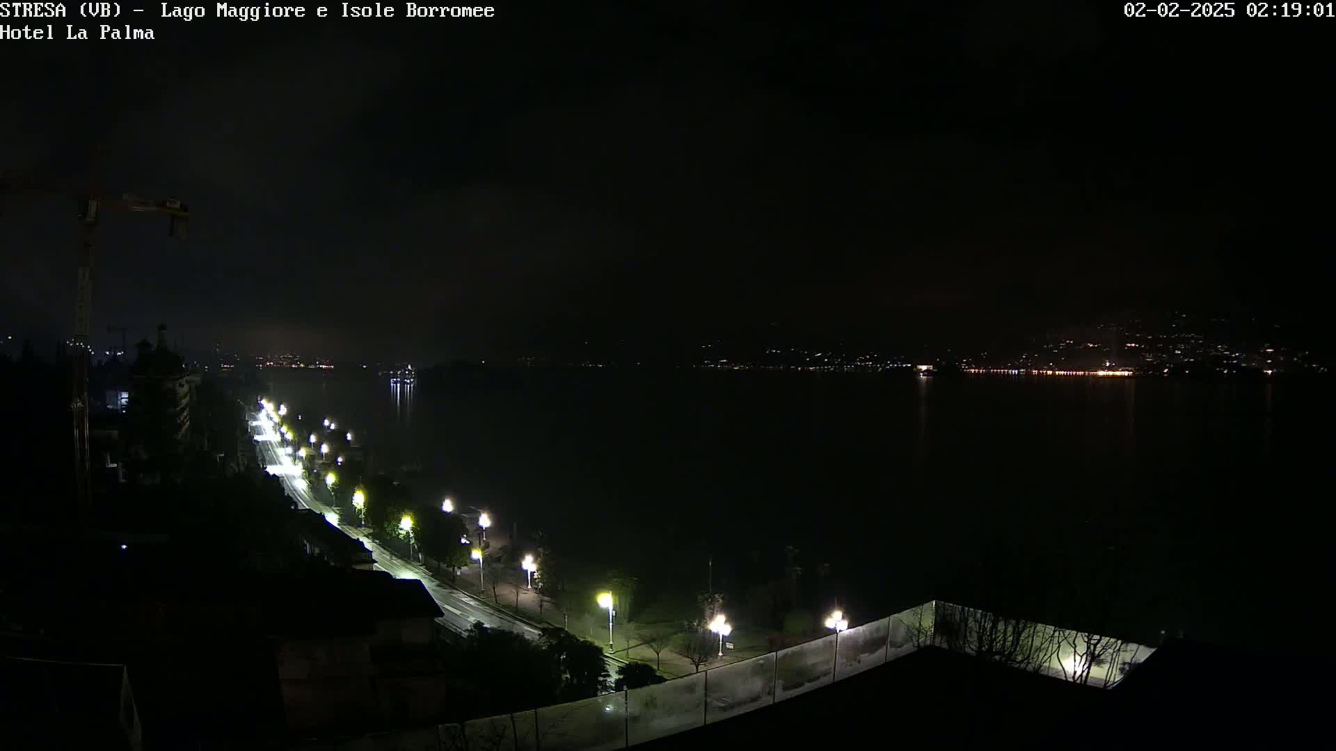 A nighttime long shot shows a dark body of water with a line of lights across the far shore, and a road with streetlights leading down to it from a higher vantage point.