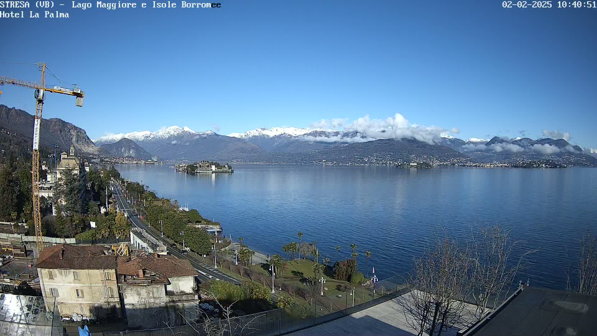 A partly cloudy, sunny day overlooking a lake with snow-capped mountains in the background, a town on the opposite shore, and a construction crane visible in the foreground.