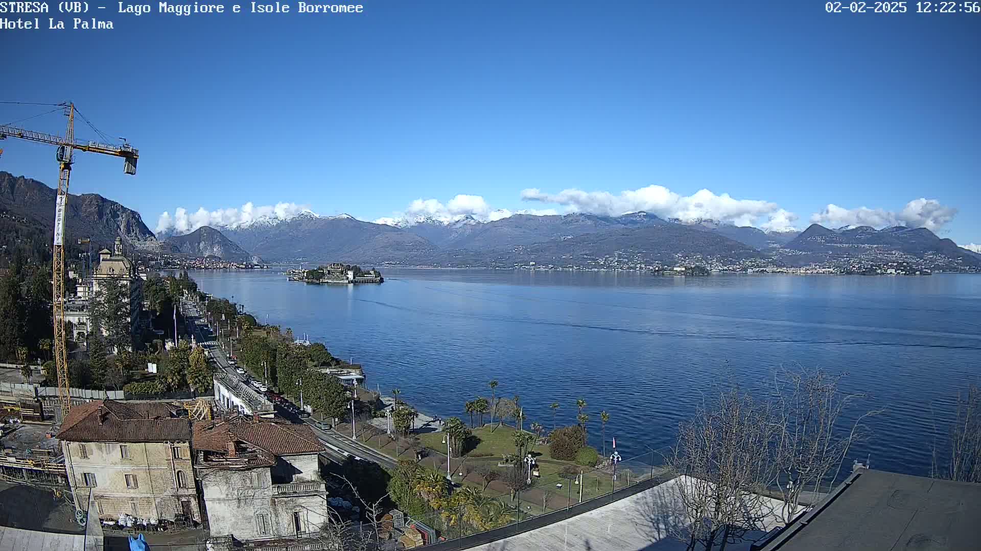 A partly sunny day shows a large lake with snow-capped mountains in the background, a small island in the middle distance, and a town along the lakeshore with a construction crane.