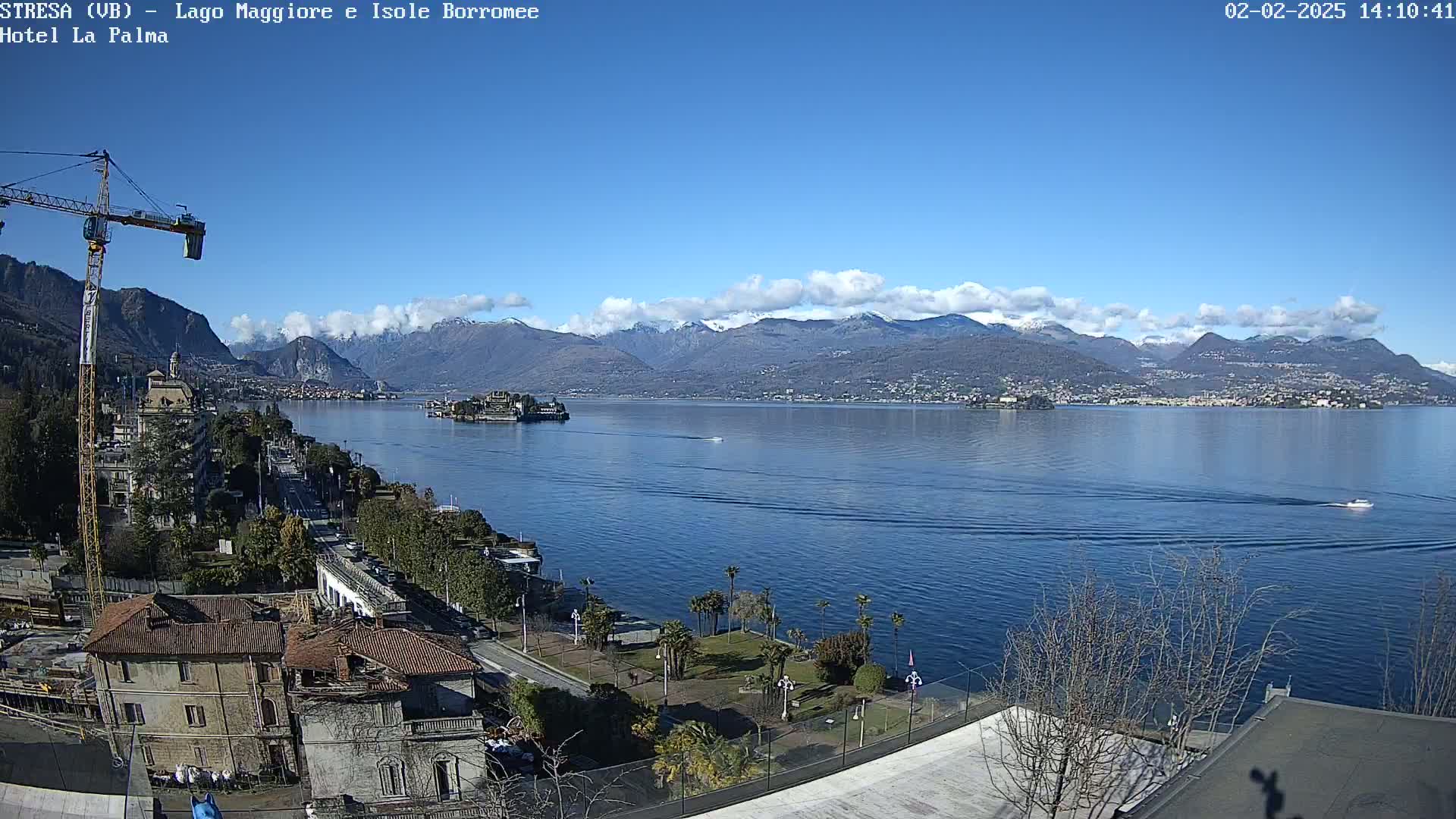 A sunny day reveals a picturesque lake view with snow-capped mountains in the background, a small island in the middle distance, and a construction crane near the foreground.