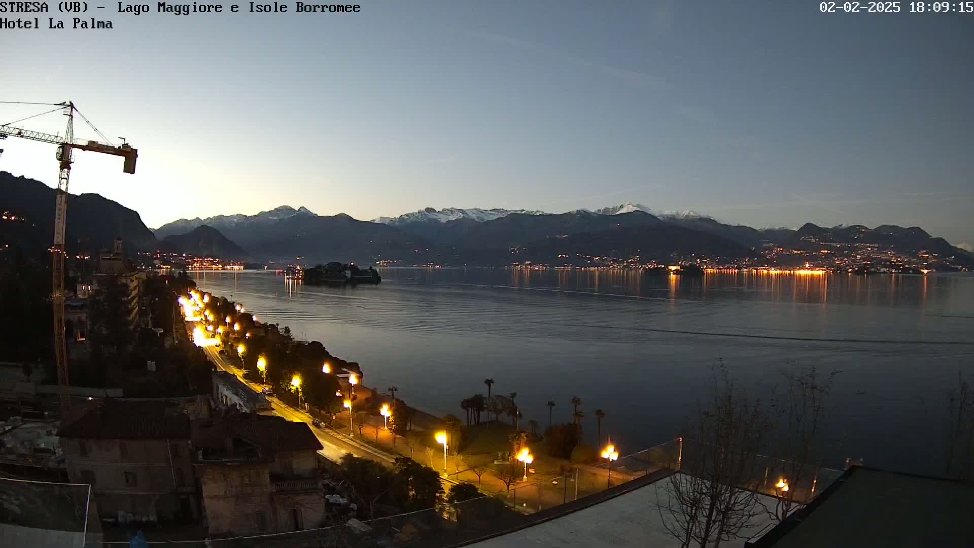 A nighttime view of a calm lake reflecting city lights and snow-capped mountains under a twilight sky, with a waterfront road illuminated by streetlights.