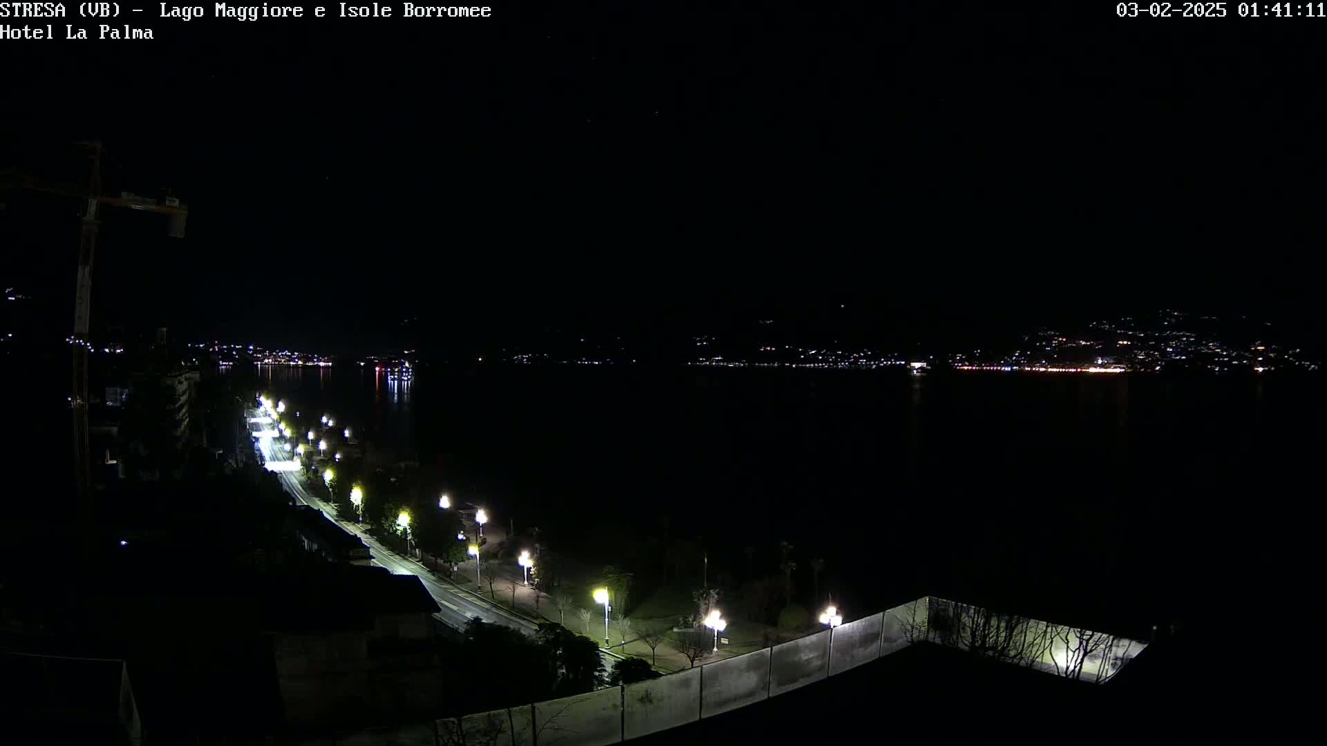A nighttime, long shot of a dimly lit road curving along a body of water with city lights visible in the distance.