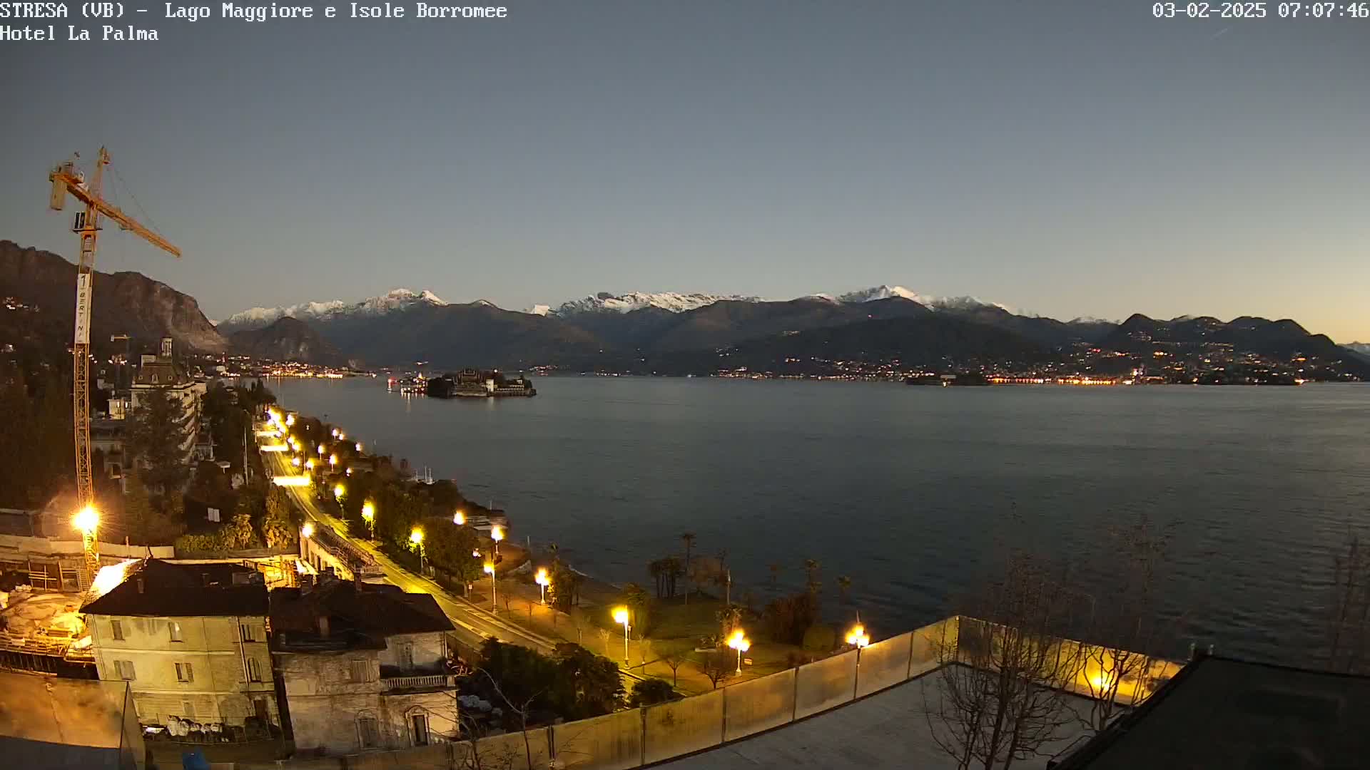 A nighttime view of a calm lake with snow-capped mountains in the background, lit by streetlights along a shoreline with a construction crane and buildings.