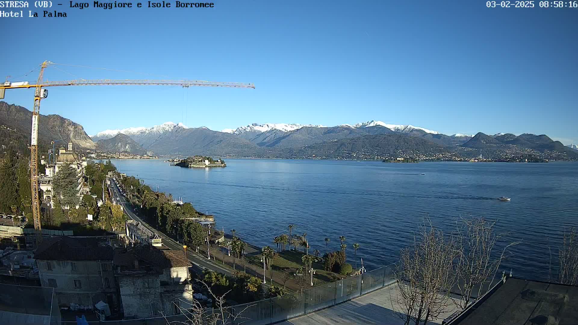 A sunny day overlooking a lake with snow-capped mountains in the background, a construction crane, a roadway, and some palm trees visible in the foreground.