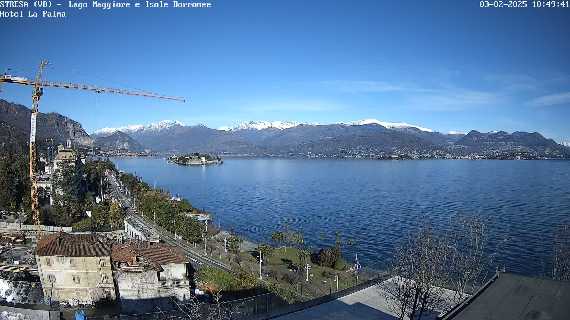 A clear, sunny day reveals a large lake with snow-capped mountains in the background, a small island in the middle of the lake, a shoreline with buildings and trees, and a construction crane near the foreground.