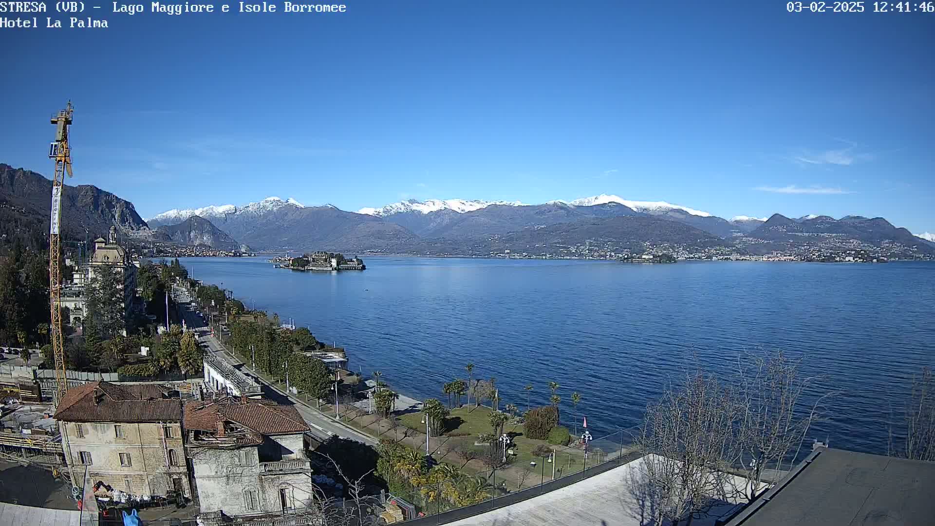 A clear, sunny day overlooks a large lake with snow-capped mountains in the background, a small island in the middle of the lake, and buildings and trees along the lakeshore.