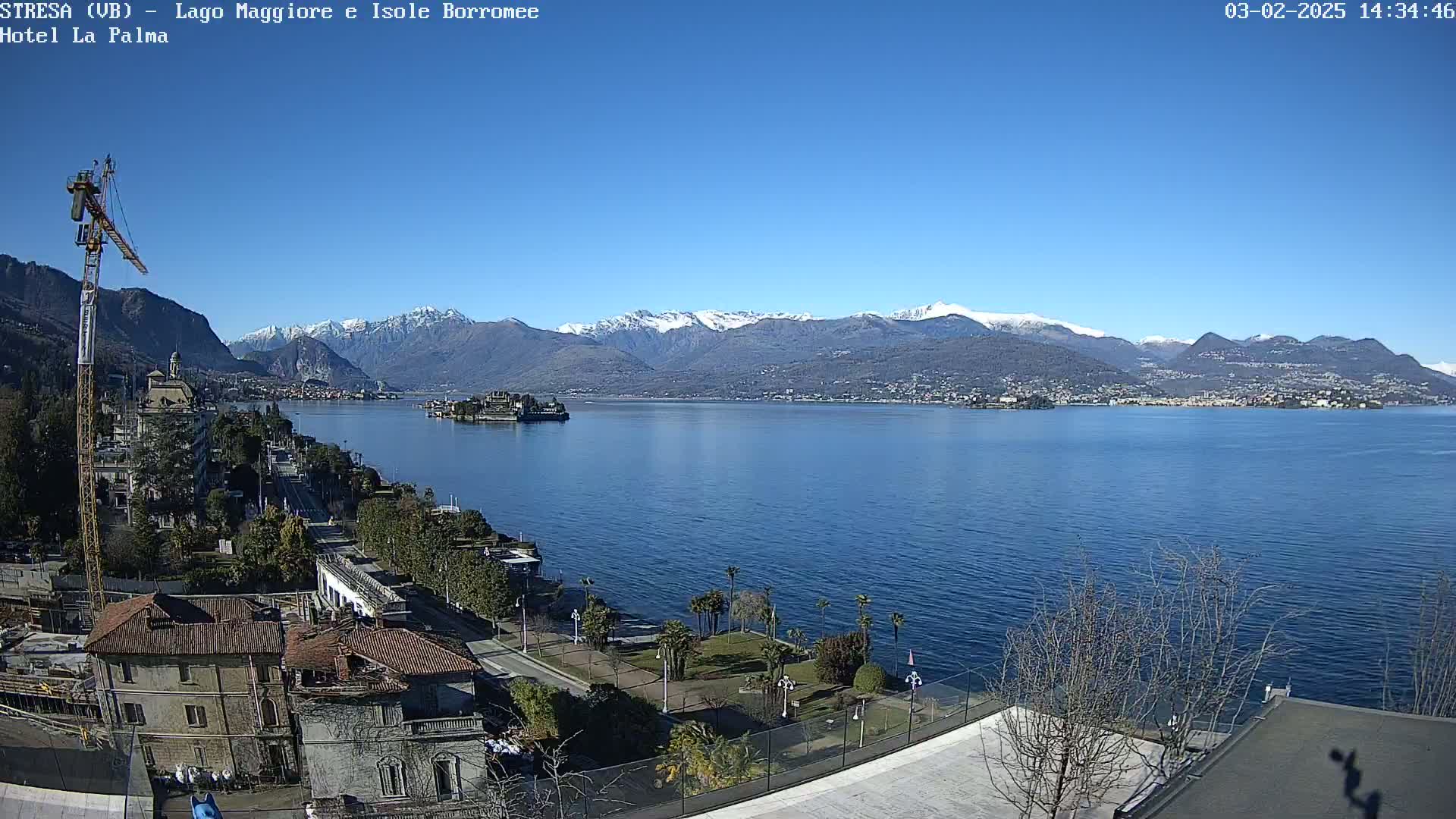 A clear, sunny day reveals a large lake with snow-capped mountains in the background, a small island in the foreground, and a town's buildings and construction crane along the lakeshore.