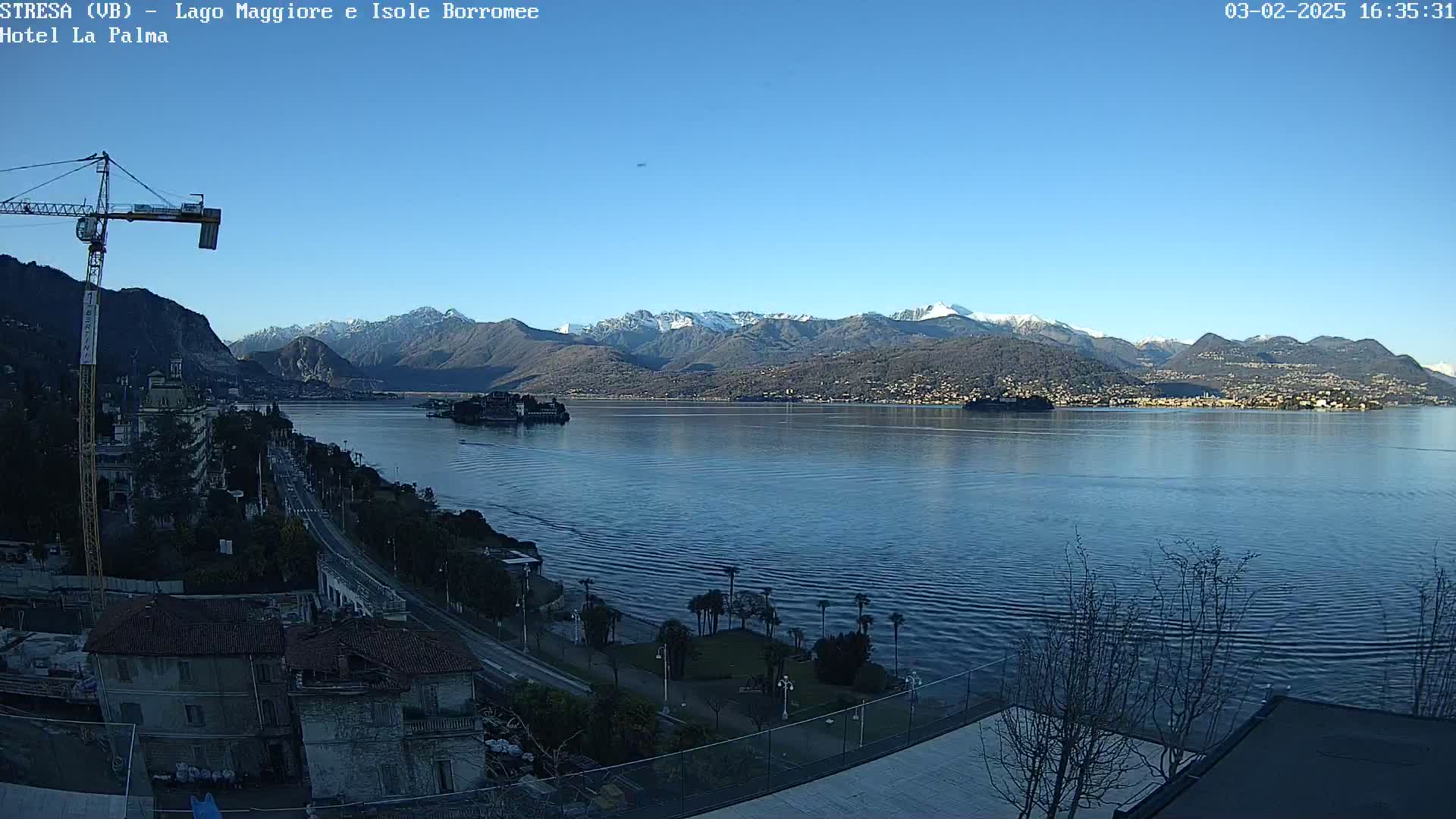 A clear, sunny day reveals a large lake surrounded by snow-capped mountains and a shoreline town, with a construction crane visible in the foreground.