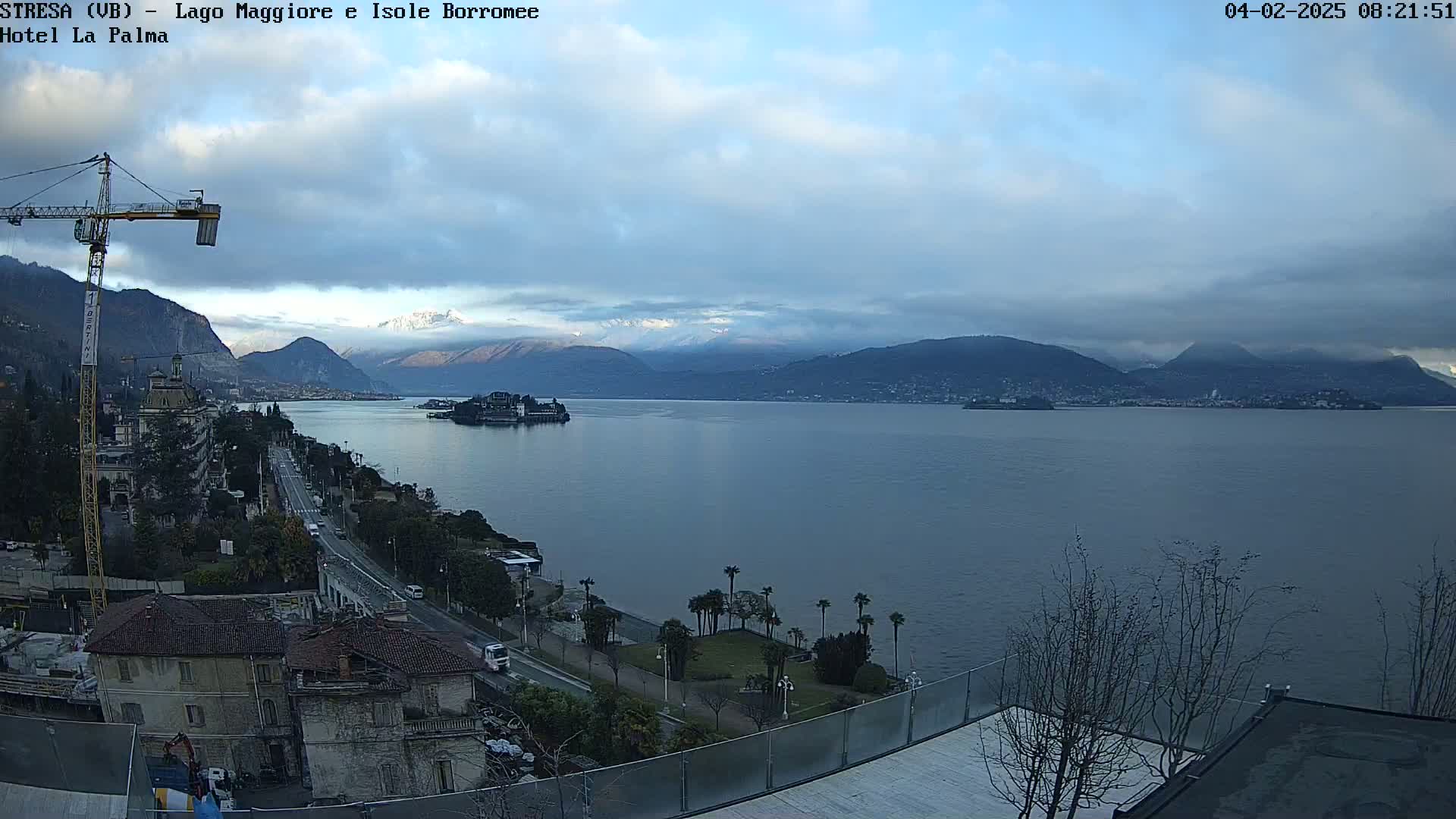 A partly cloudy day overlooks a large body of water with snow-capped mountains in the distance, a shoreline with buildings and trees, and a construction crane visible near the foreground.