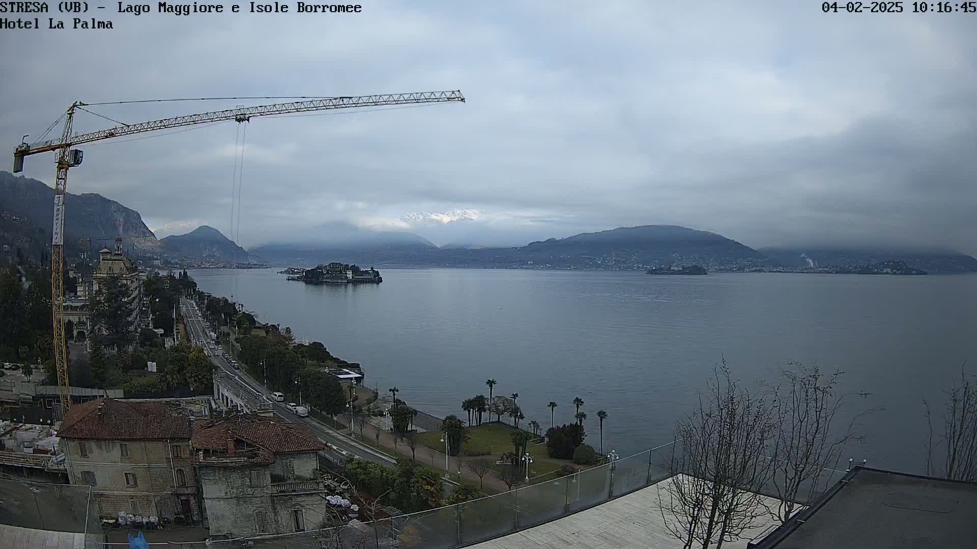 A large body of water is seen from an elevated vantage point, under an overcast sky, with a crane, buildings, and trees in the foreground, and mountains in the distance.