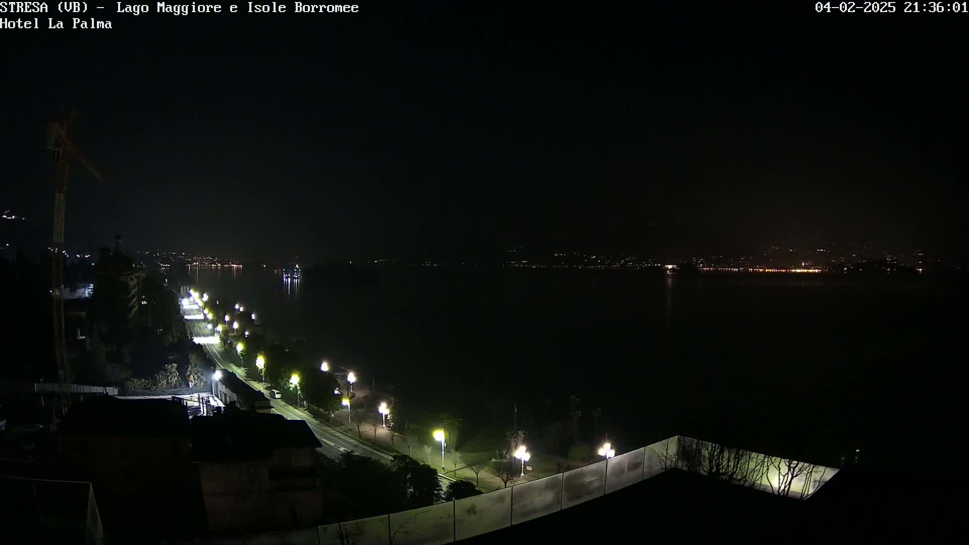 A nighttime view of a lakeside road with illuminated streetlights, overlooking a dark body of water with faintly visible lights on the opposite shore under a clear, dark sky.