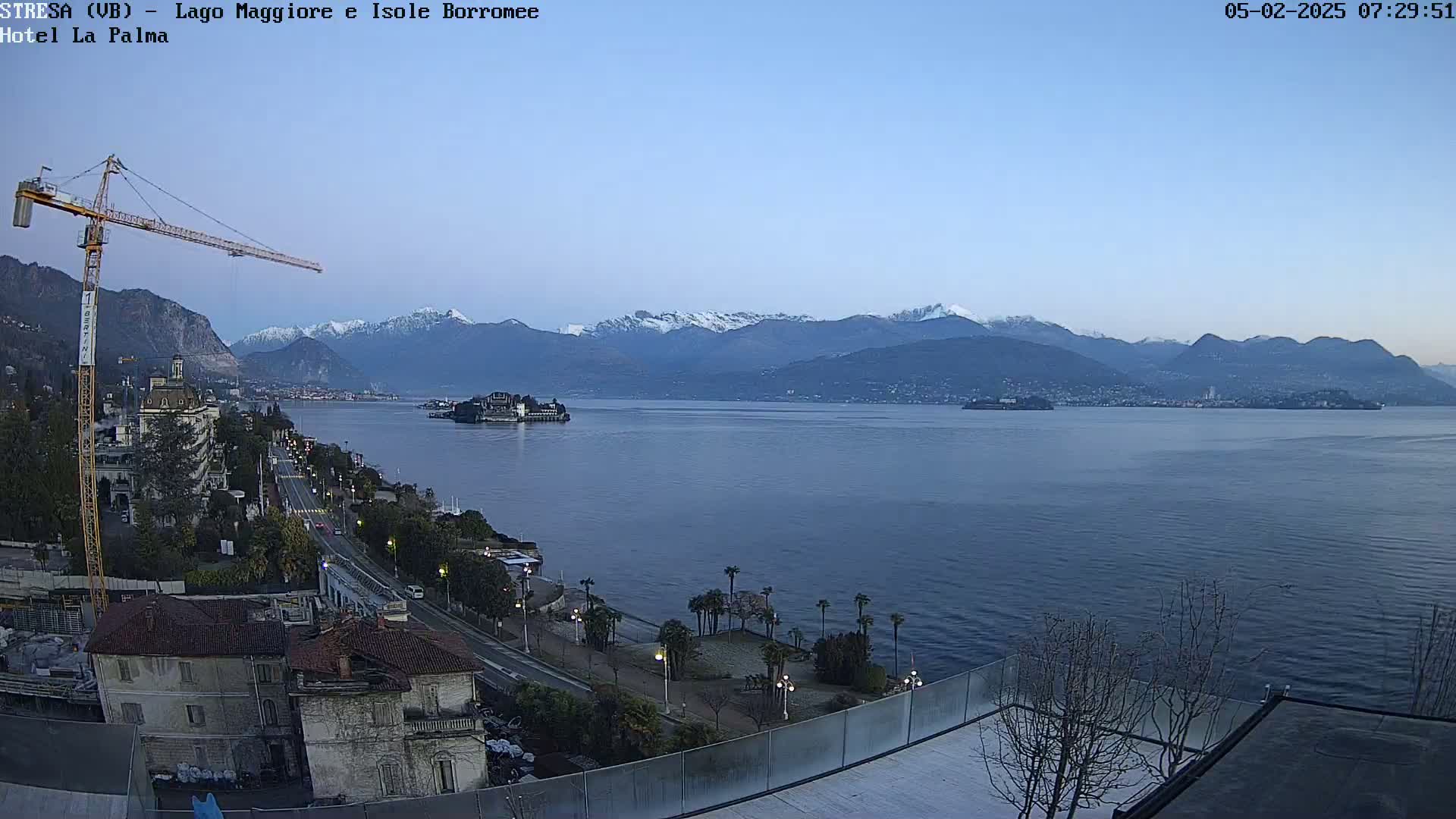 A tranquil lake scene at dawn, featuring snow-capped mountains in the background, a small island in the middle of the lake, and buildings along the lakeshore.