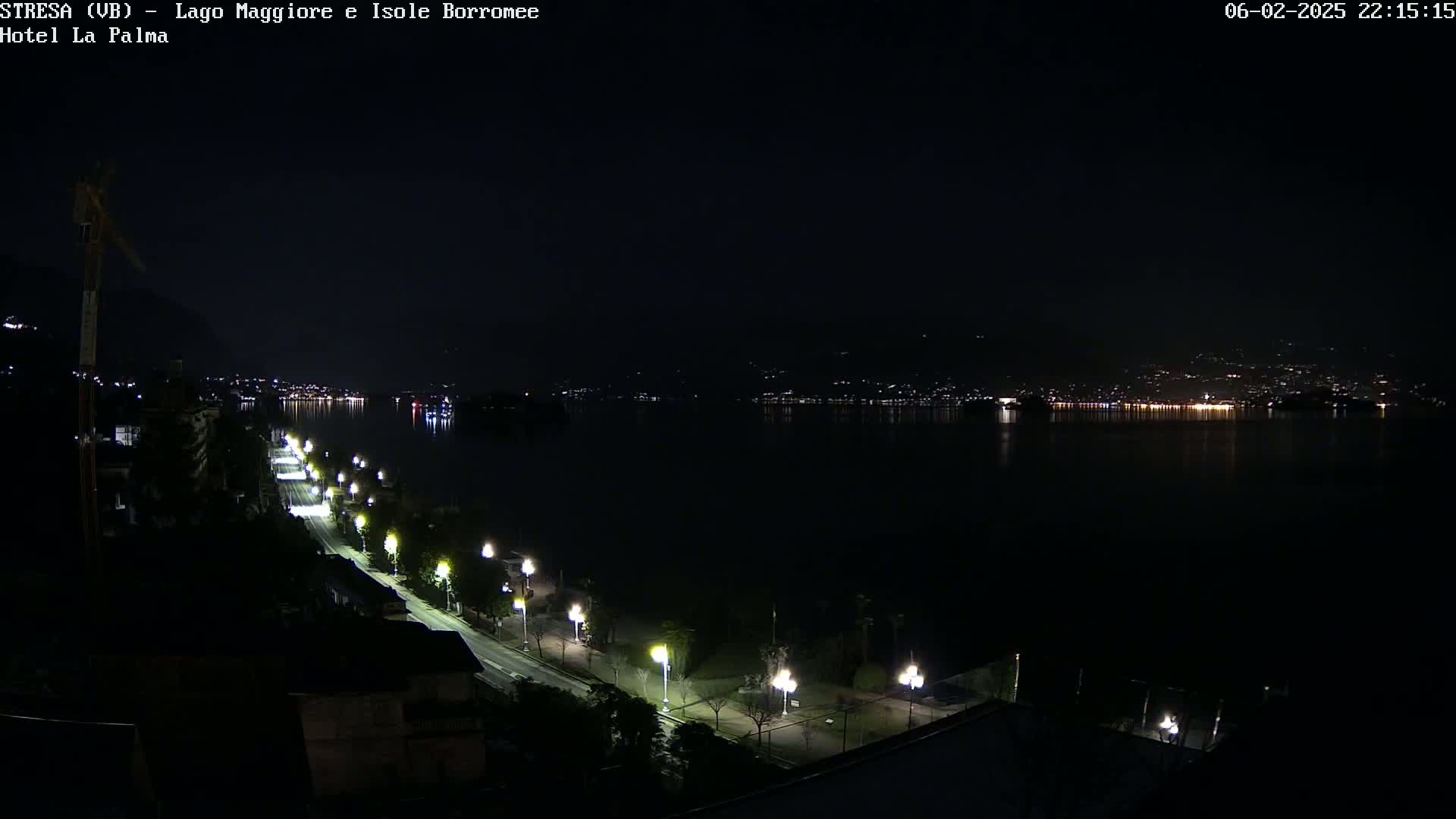 A nighttime view of a dark, calm lake with a shoreline road lined with illuminated streetlights, and distant city lights reflected in the water.