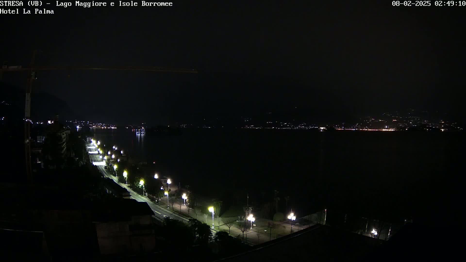 A nighttime view of a dark, calm lake with a city's lights visible in the distance, and a road lined with lit street lamps curving along the lakeshore.