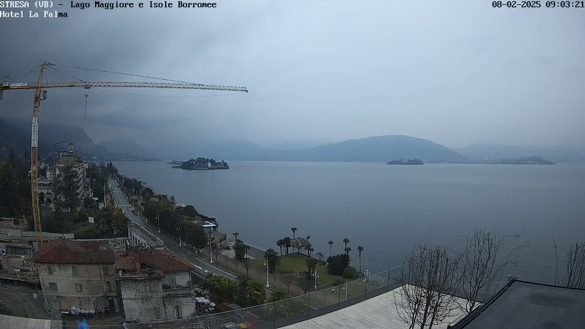 A large body of water is seen under an overcast sky, with a shoreline featuring buildings, a road, vegetation, and a construction crane.