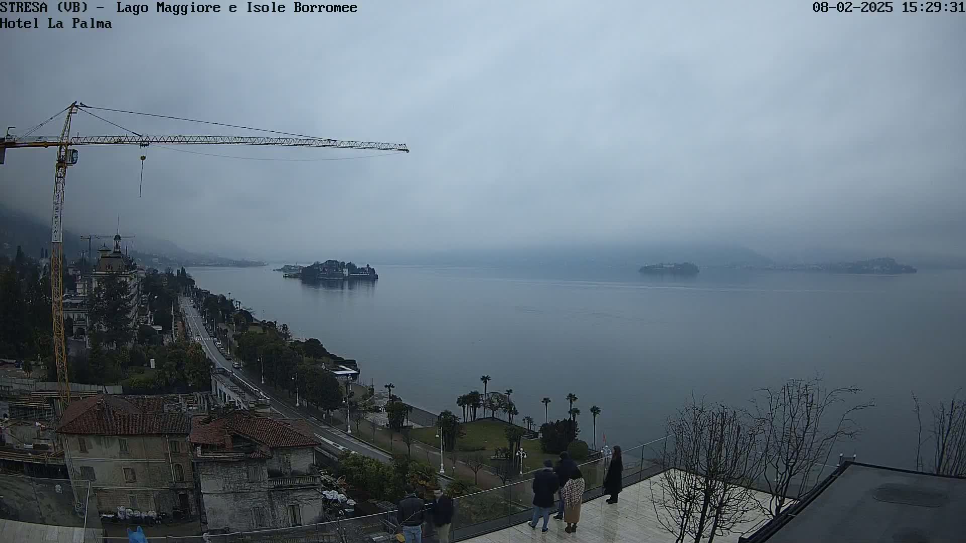 A hazy, overcast day reveals a large body of water with islands in the distance, a town situated along the shoreline with a construction crane present, and several people standing on a rooftop patio.