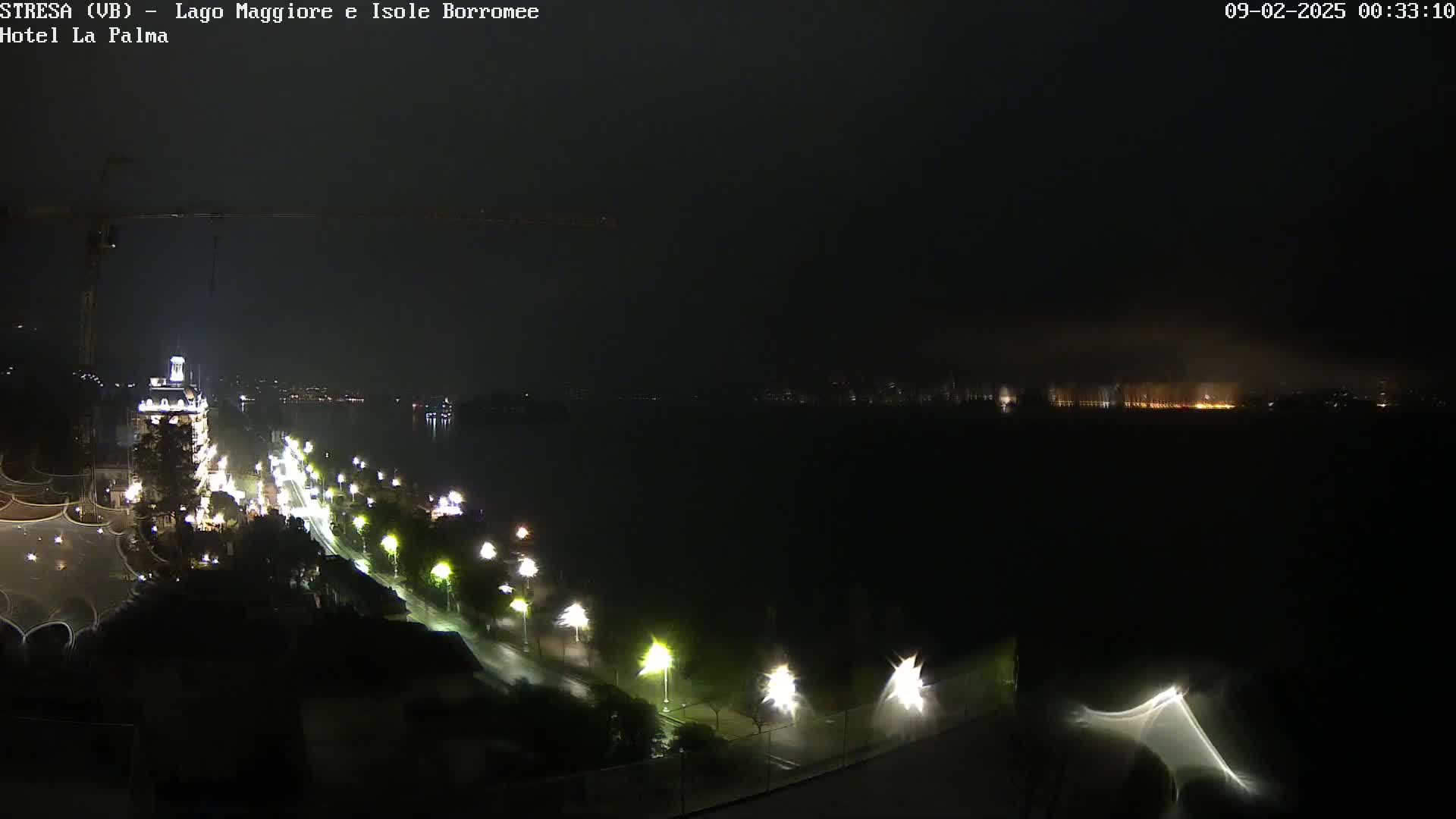 A nighttime view of a city and lake illuminated by streetlights and distant lights across the water, under a dark, possibly overcast sky.