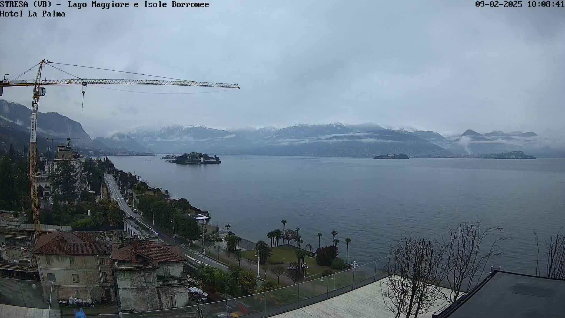 A large body of water is seen with snow-capped mountains in the background under an overcast sky, a construction crane and buildings along the shore are visible in the foreground.