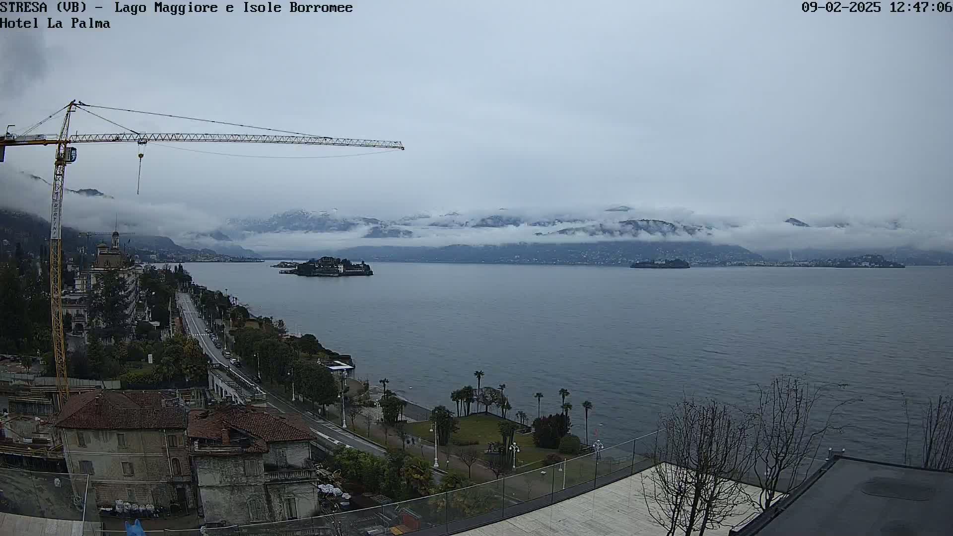 A large body of water stretches out before a town nestled against snow-capped mountains partially obscured by clouds under an overcast sky, a construction crane visible to the left.