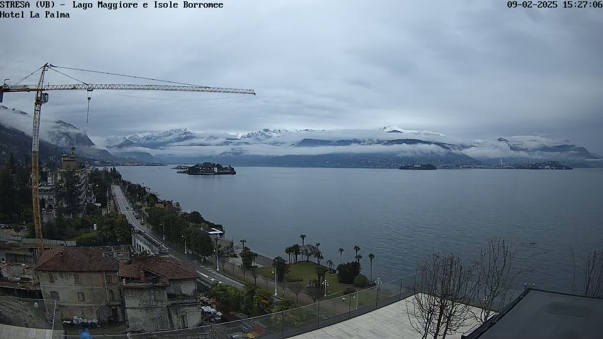 A large body of water stretches toward snow-capped mountains partially obscured by fog, under a cloudy sky, with a crane visible near buildings and landscaping in the foreground.