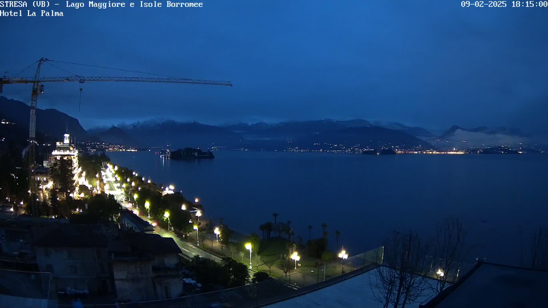 A nighttime view of a lake with a city and snow-capped mountains in the background, under a twilight sky; a construction crane is visible in the foreground.
