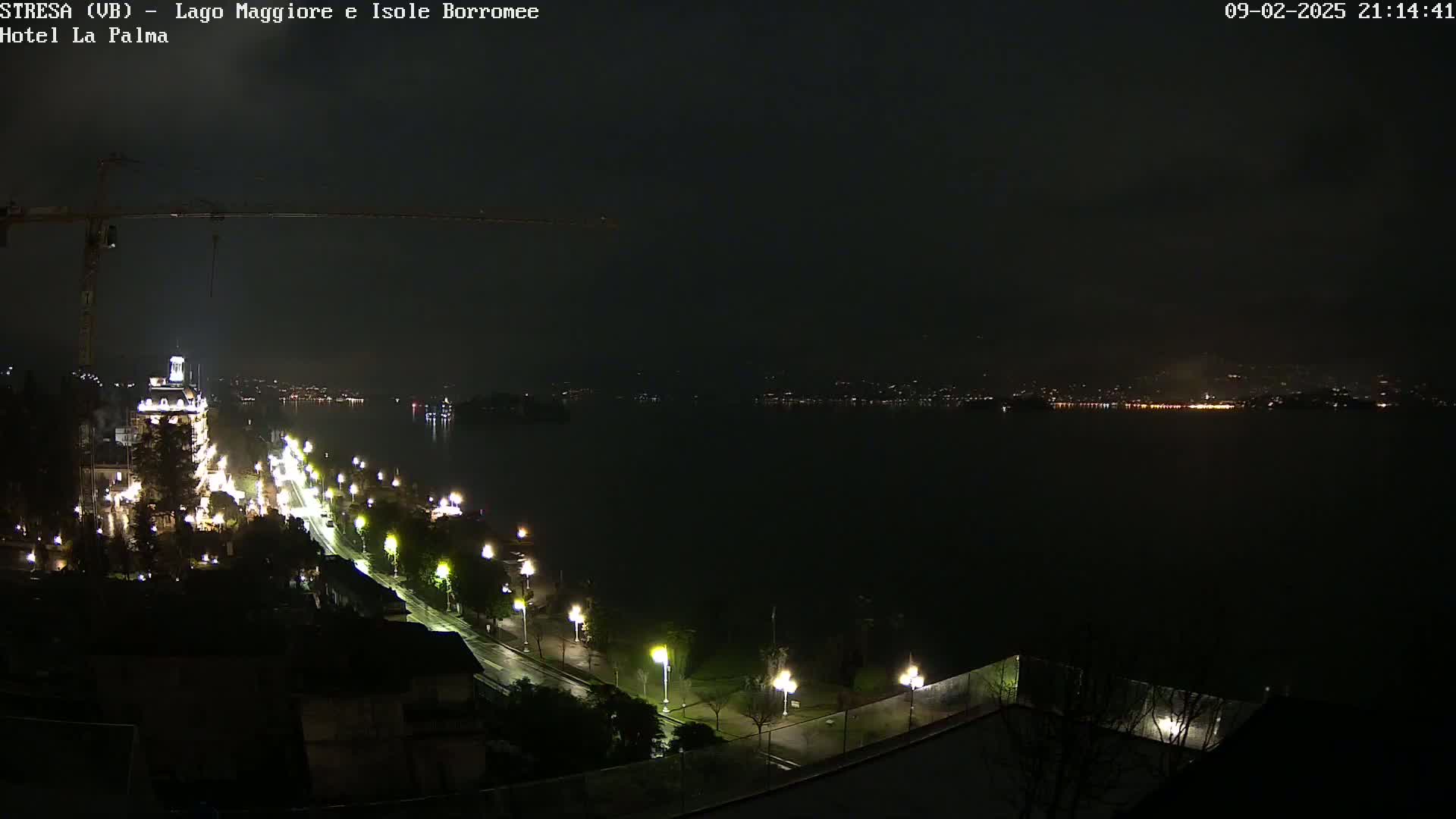 A nighttime long shot of a city waterfront illuminated by streetlights, with a construction crane visible on the left and a dark body of water extending to a distant, faintly lit shoreline under a cloudy sky.