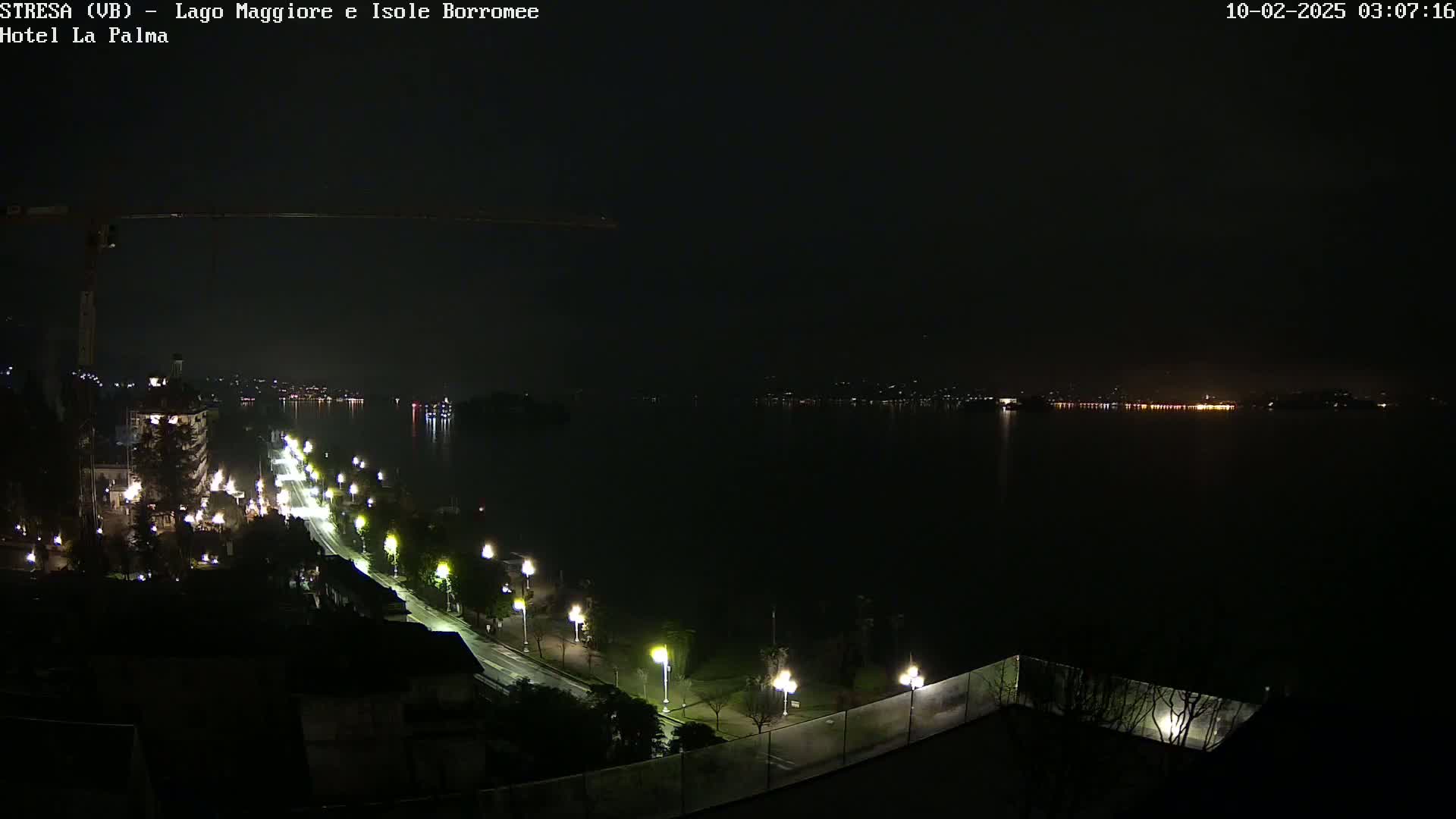 A nighttime view of a city street illuminated by streetlights along a body of water with a distant shoreline faintly visible.
