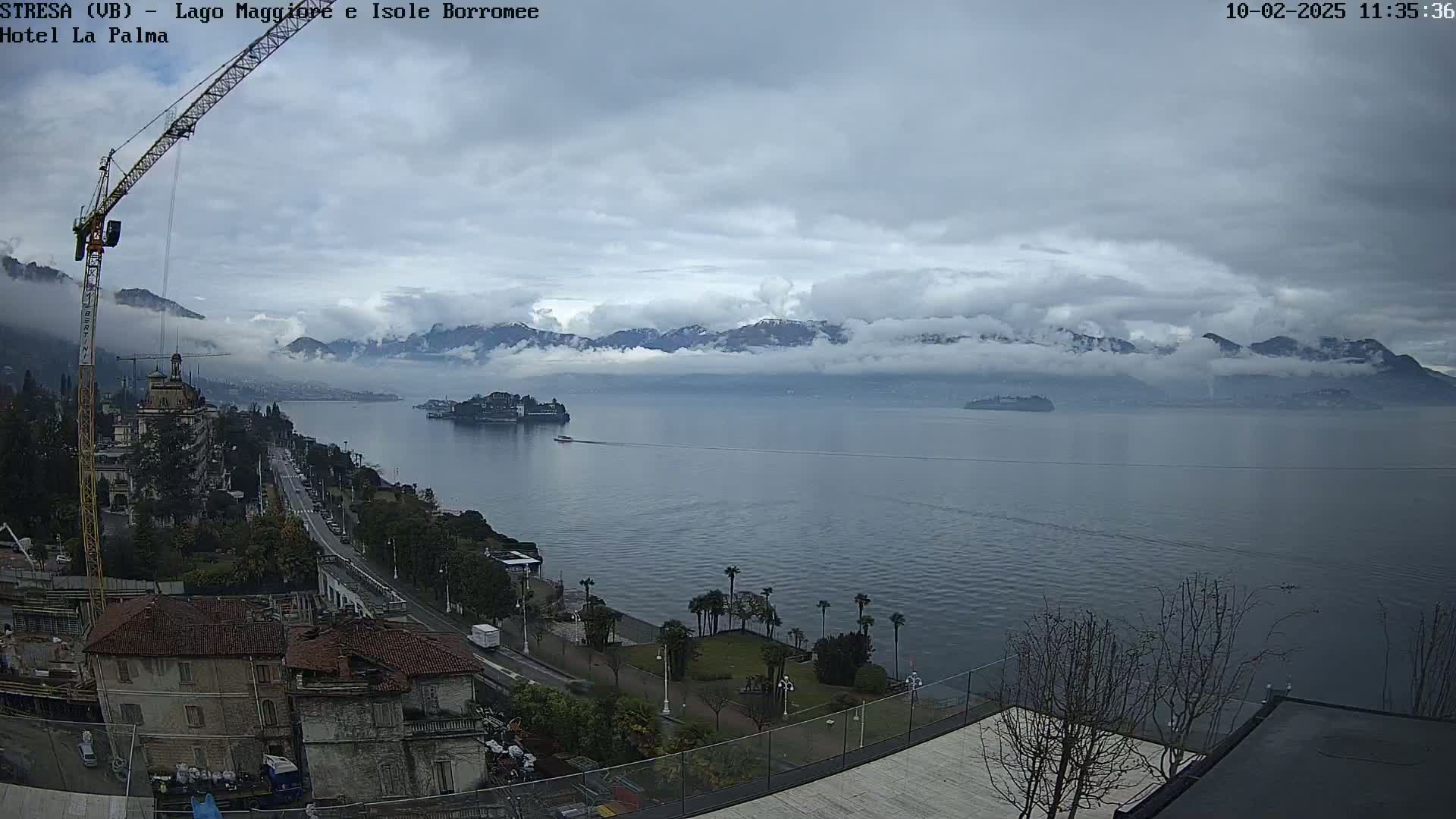 A large body of water stretches out under a cloudy sky toward distant mountains partially obscured by fog, with a crane visible near buildings on the shoreline.