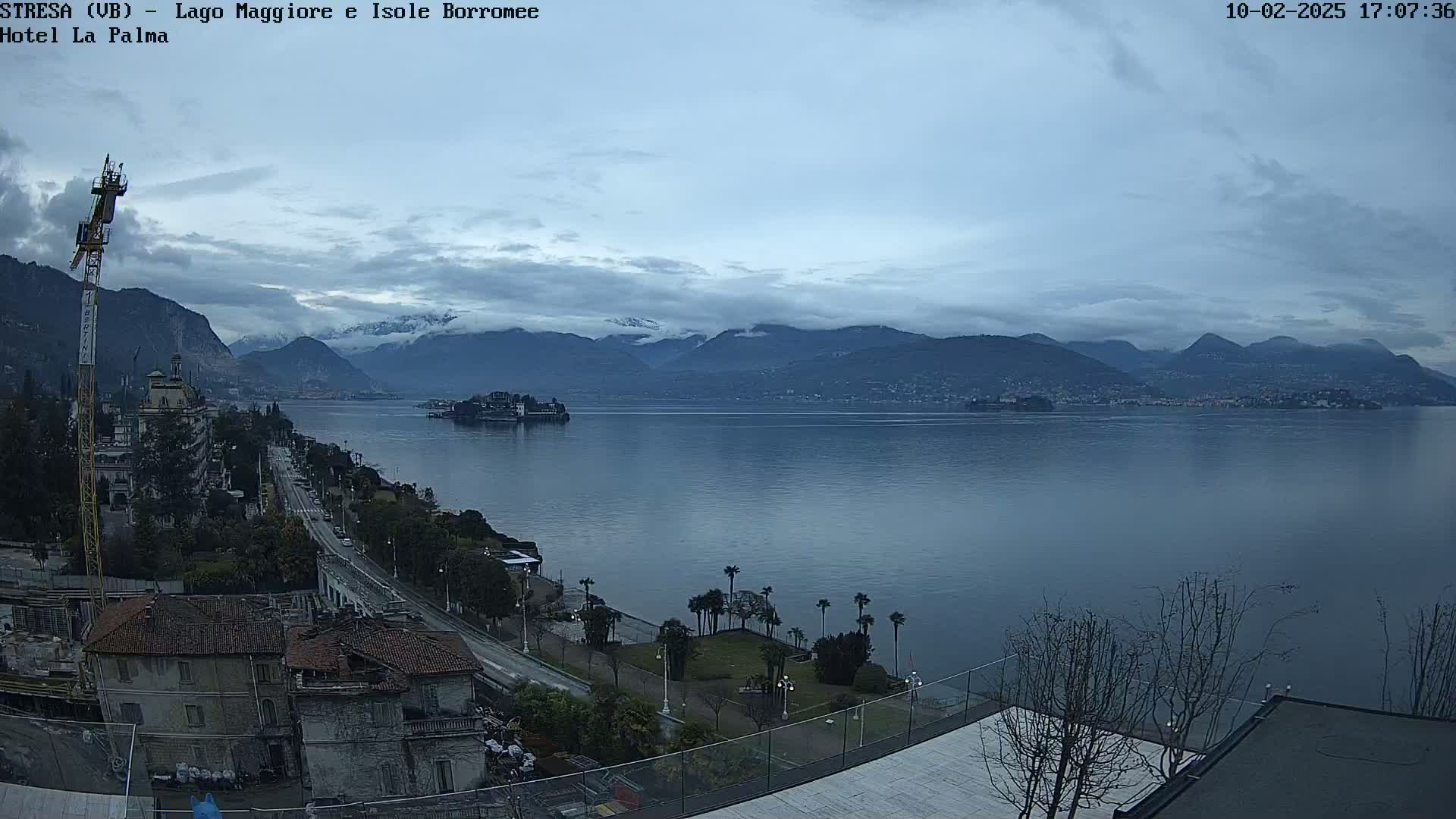A lake stretches out before snow-capped mountains under an overcast sky, with a town and construction site visible in the foreground.