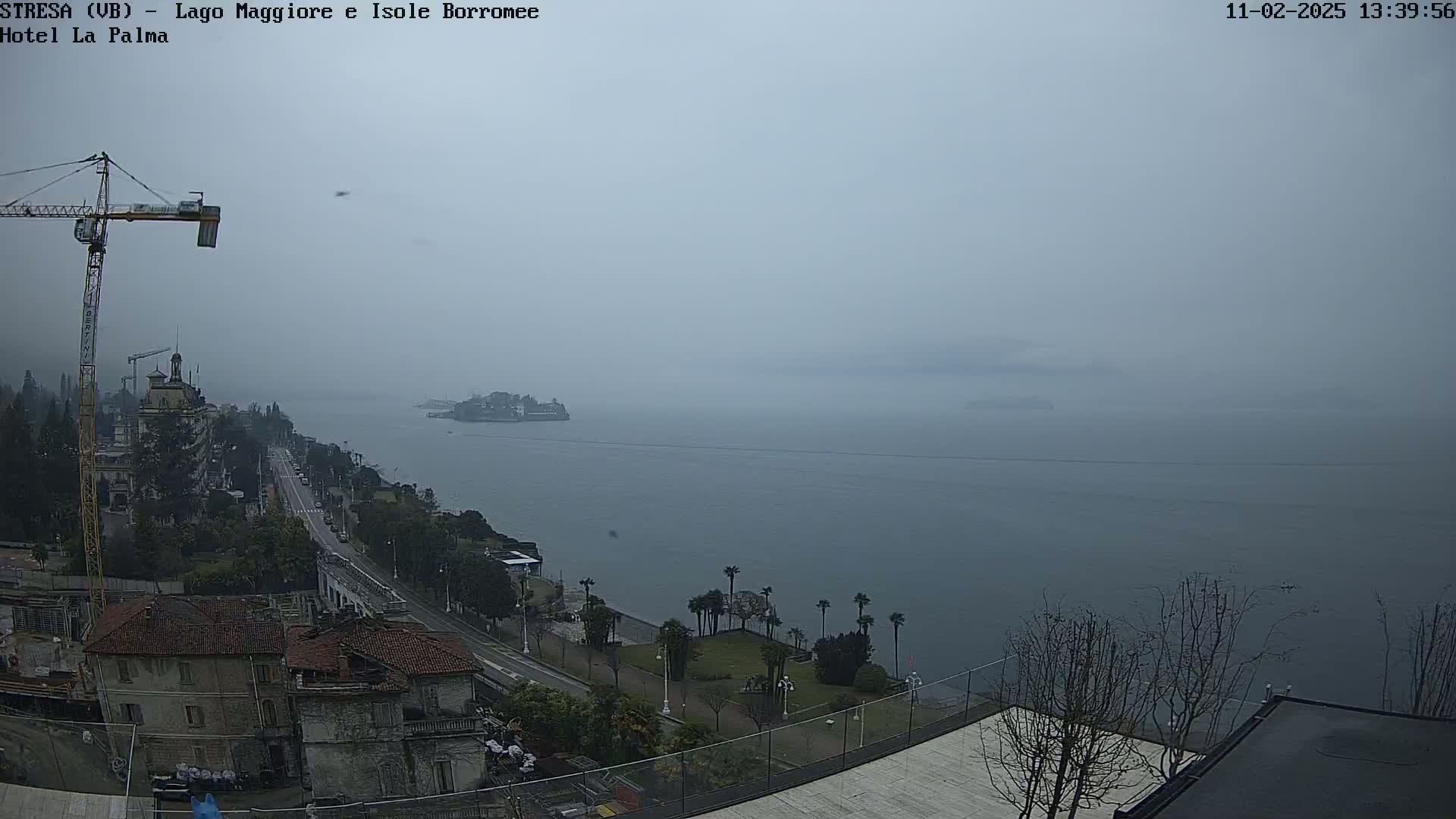 A hazy, overcast day reveals a coastal town with a construction crane, a road leading to a calm body of water, and distant islands shrouded in mist.