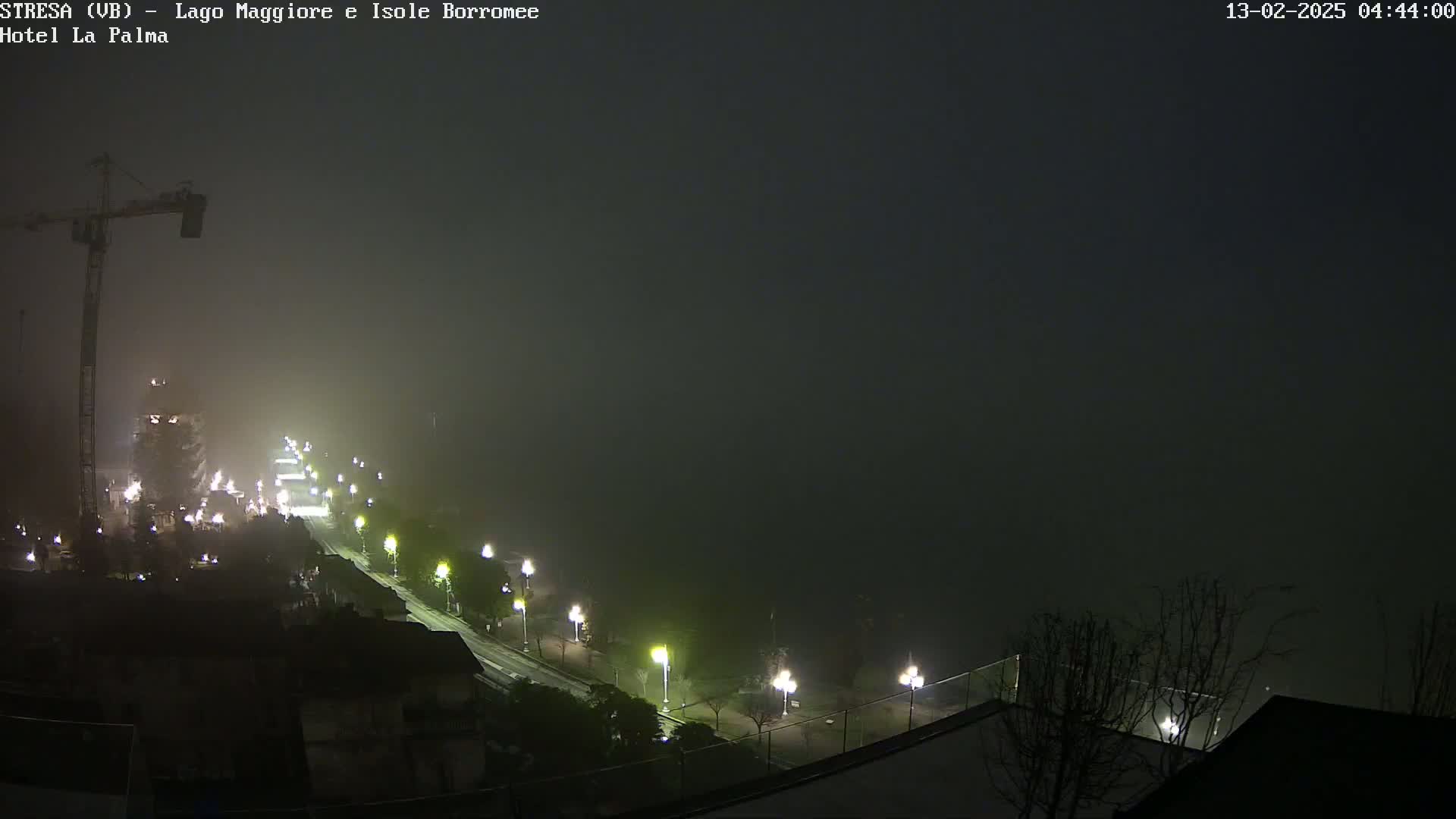 A foggy night scene shows a road with lights leading to a body of water, a construction crane, and buildings along the roadside.