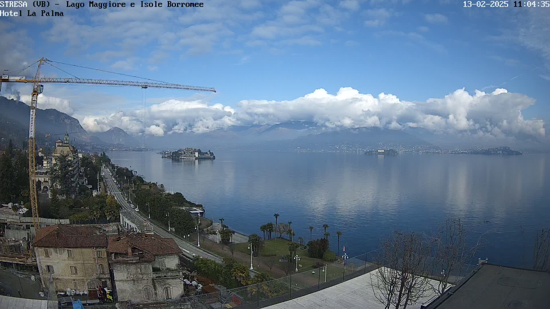 A large body of water with mountains in the background, under a partly cloudy sky, is viewed from a high vantage point showing buildings, a road, and a construction crane.
