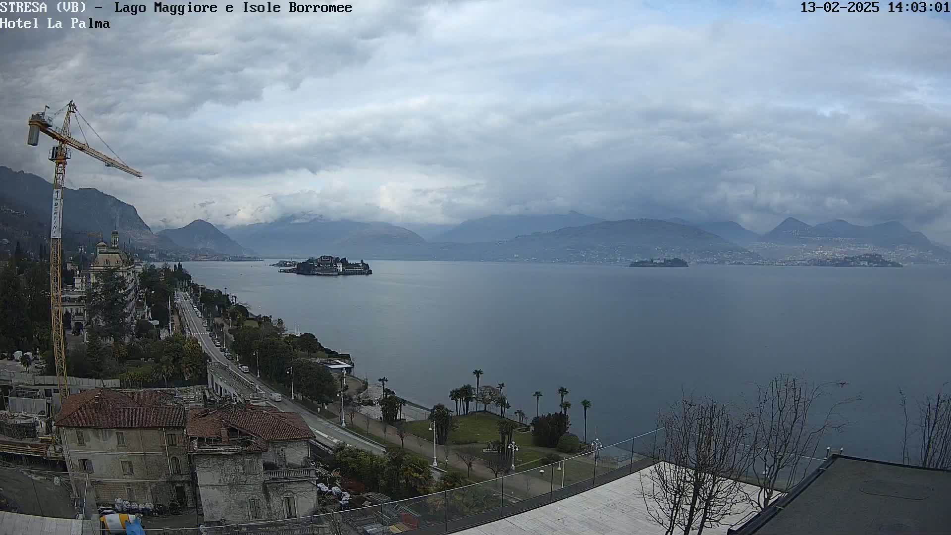 A large body of water is seen under an overcast sky, with mountains in the distance, a shoreline with buildings and trees, and a construction crane.