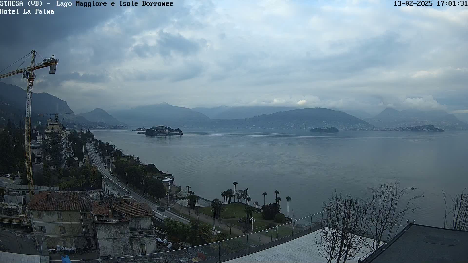 A large lake is seen under a cloudy sky, with mountains in the background and a town visible along the shoreline, including a construction crane and palm trees.