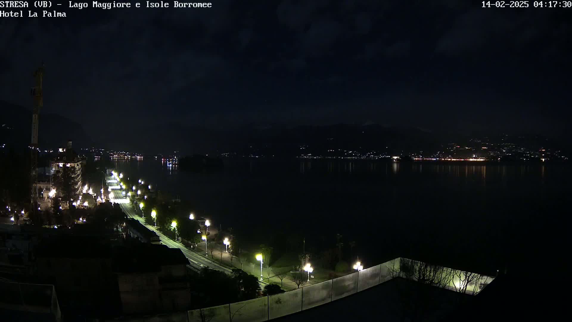 A nighttime, dimly lit lakeside road with sparsely spaced streetlights curves along a dark body of water, with faintly visible lights across the water and hills in the distance under a mostly cloudy sky.
