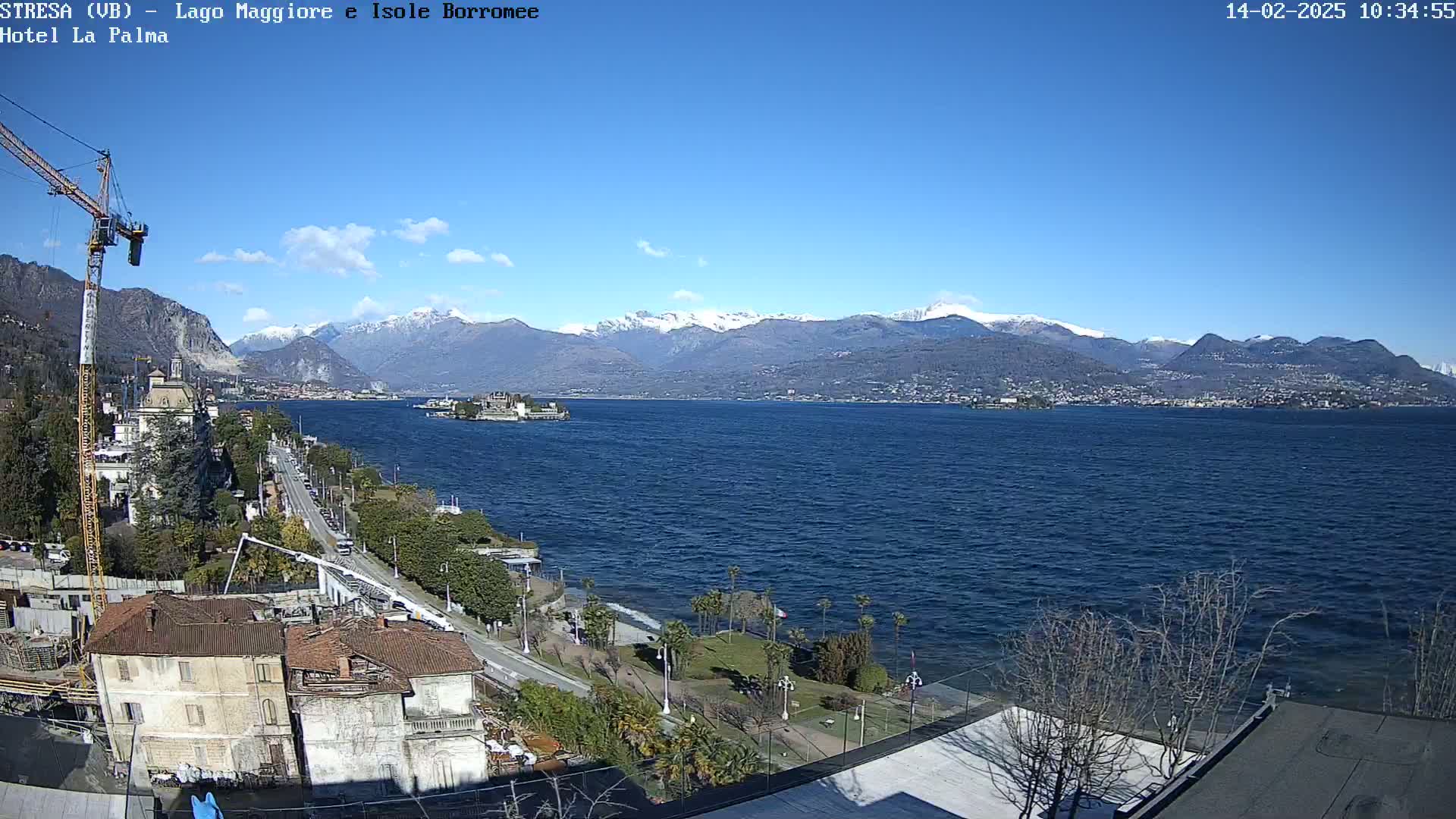A sunny day reveals a large lake with snow-capped mountains in the background, a small island in the middle of the lake, and a lakeside town with a construction crane visible.