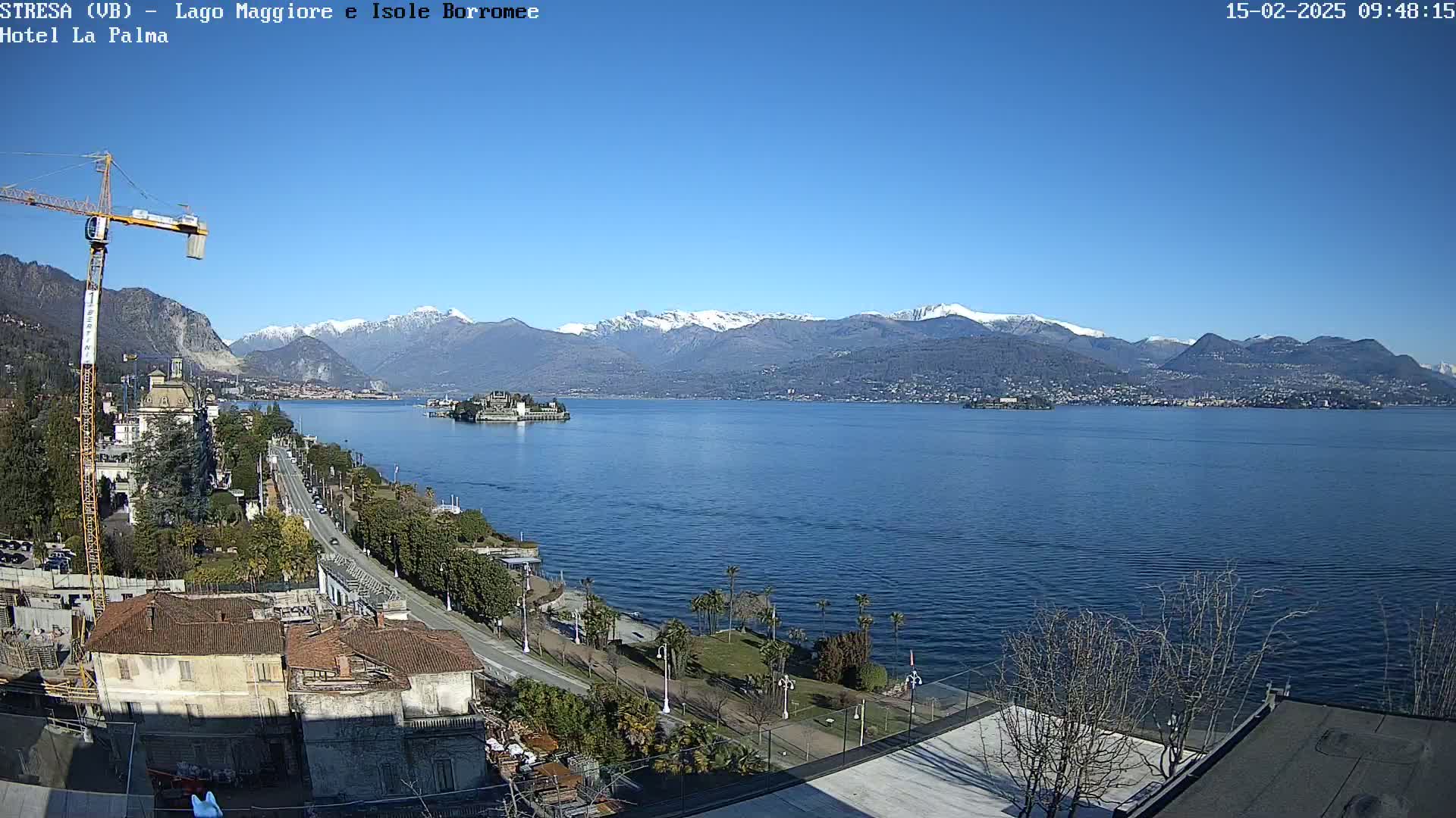 A clear, sunny day reveals a large lake, with snow-capped mountains in the background, a small island in the middle of the lake, and a waterfront road lined with buildings and trees in the foreground.