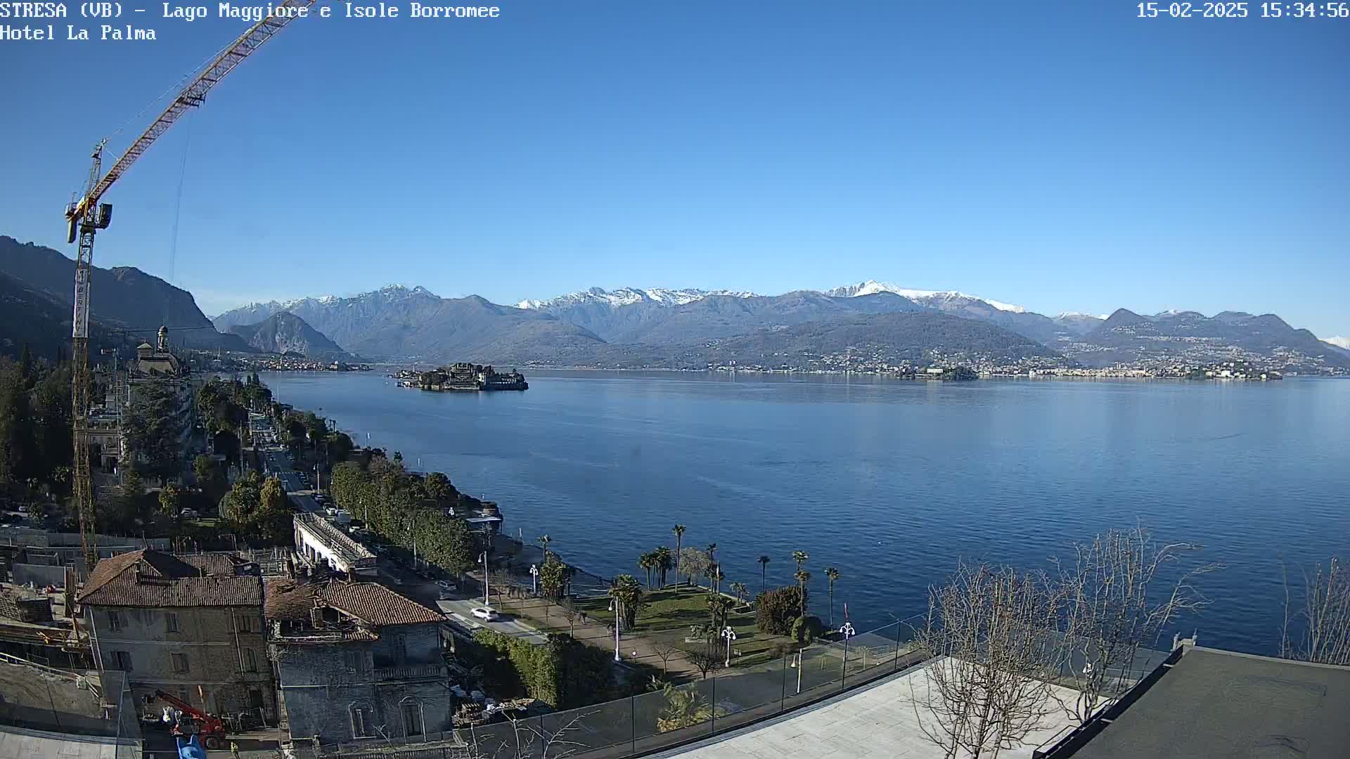 A clear, sunny day overlooks a lake with snow-capped mountains in the background, a town on the lakeshore, and a construction crane visible in the foreground.