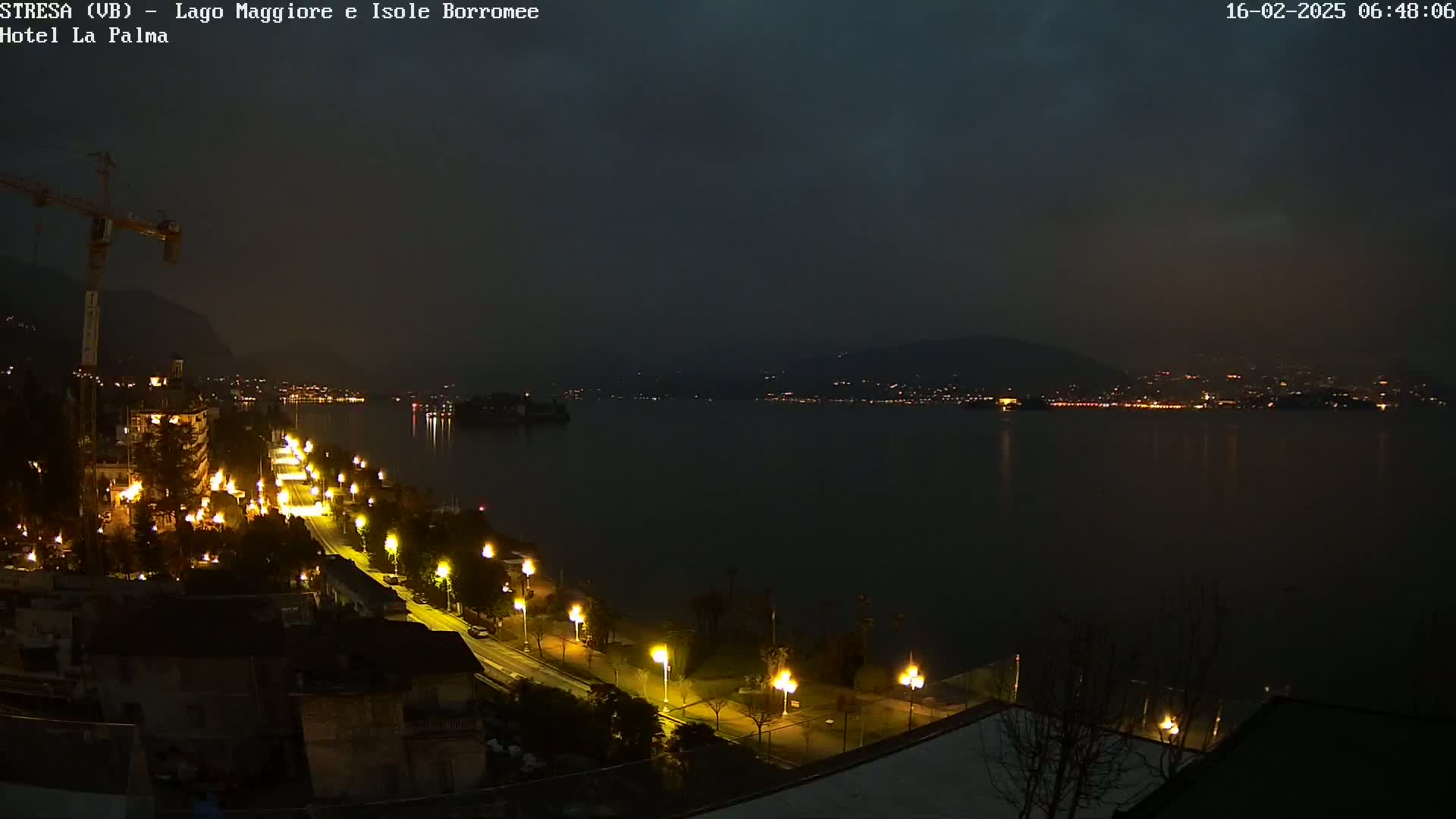 A nighttime view of a lakeside town with illuminated streets and buildings, overlooking a dark body of water under a cloudy sky.