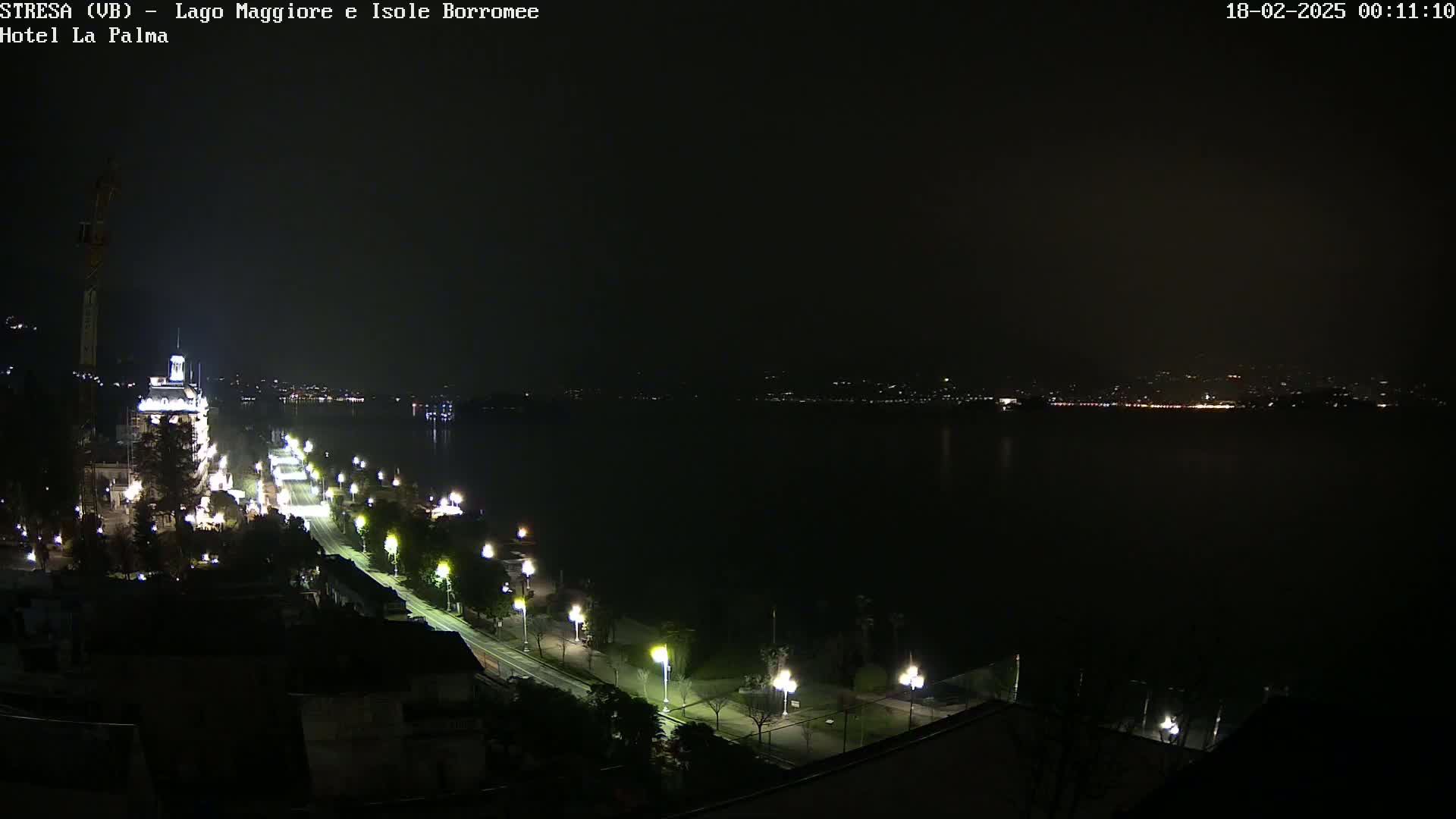 A nighttime, aerial view shows a long, illuminated road curving along a dark body of water, with faintly lit buildings visible across the water.