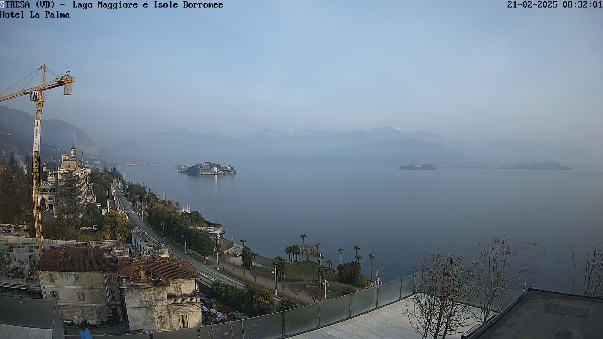 A hazy, long-range view of a lake with snow-capped mountains in the background, featuring a shoreline with buildings, a road, and a construction crane.