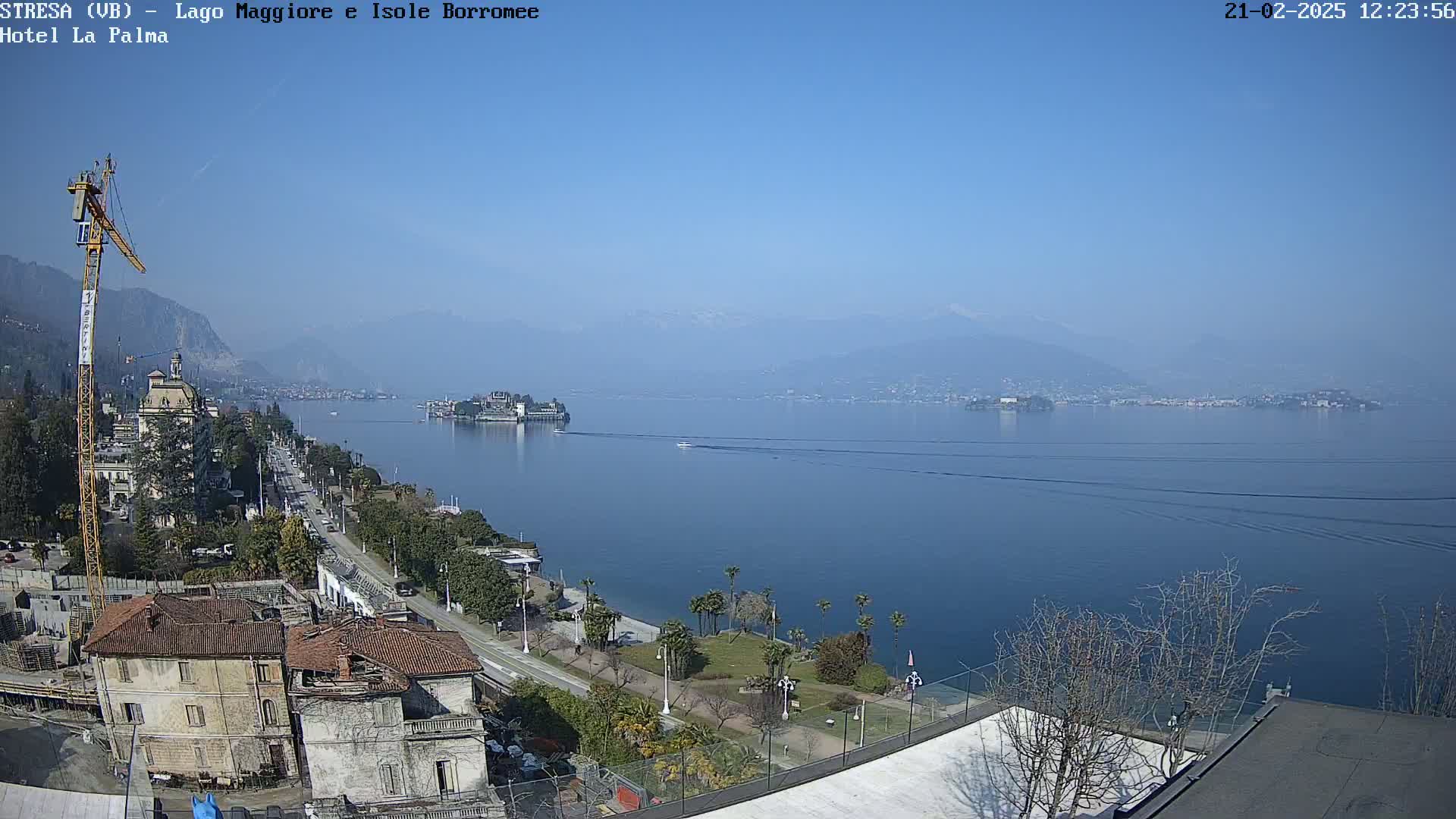 A clear, sunny day overlooks a calm lake with a shoreline featuring buildings, trees, and a small boat; mountains are visible in the distance.