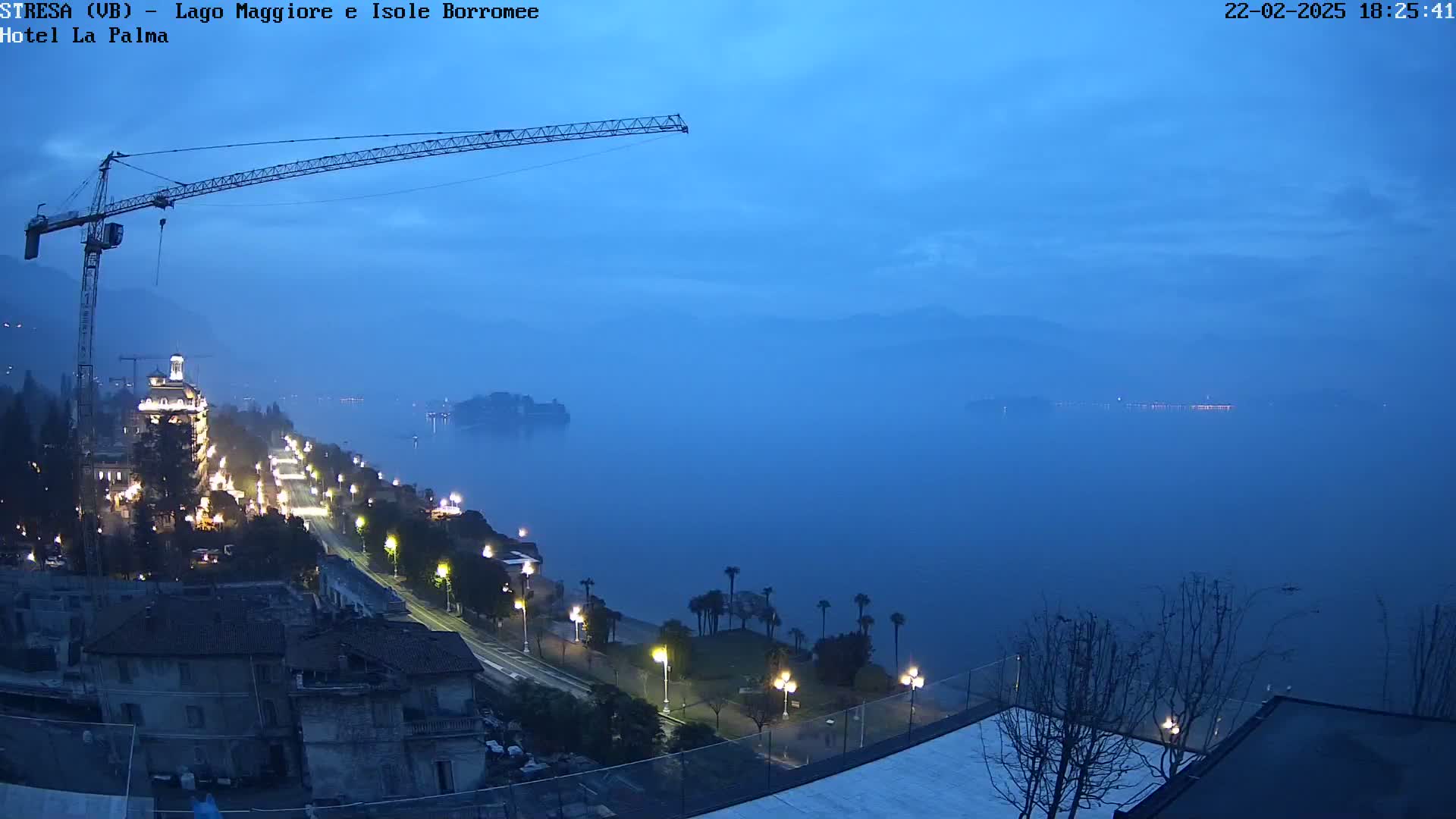 A twilight view of a lakeside town with a construction crane, illuminated buildings, and a calm, hazy lake.