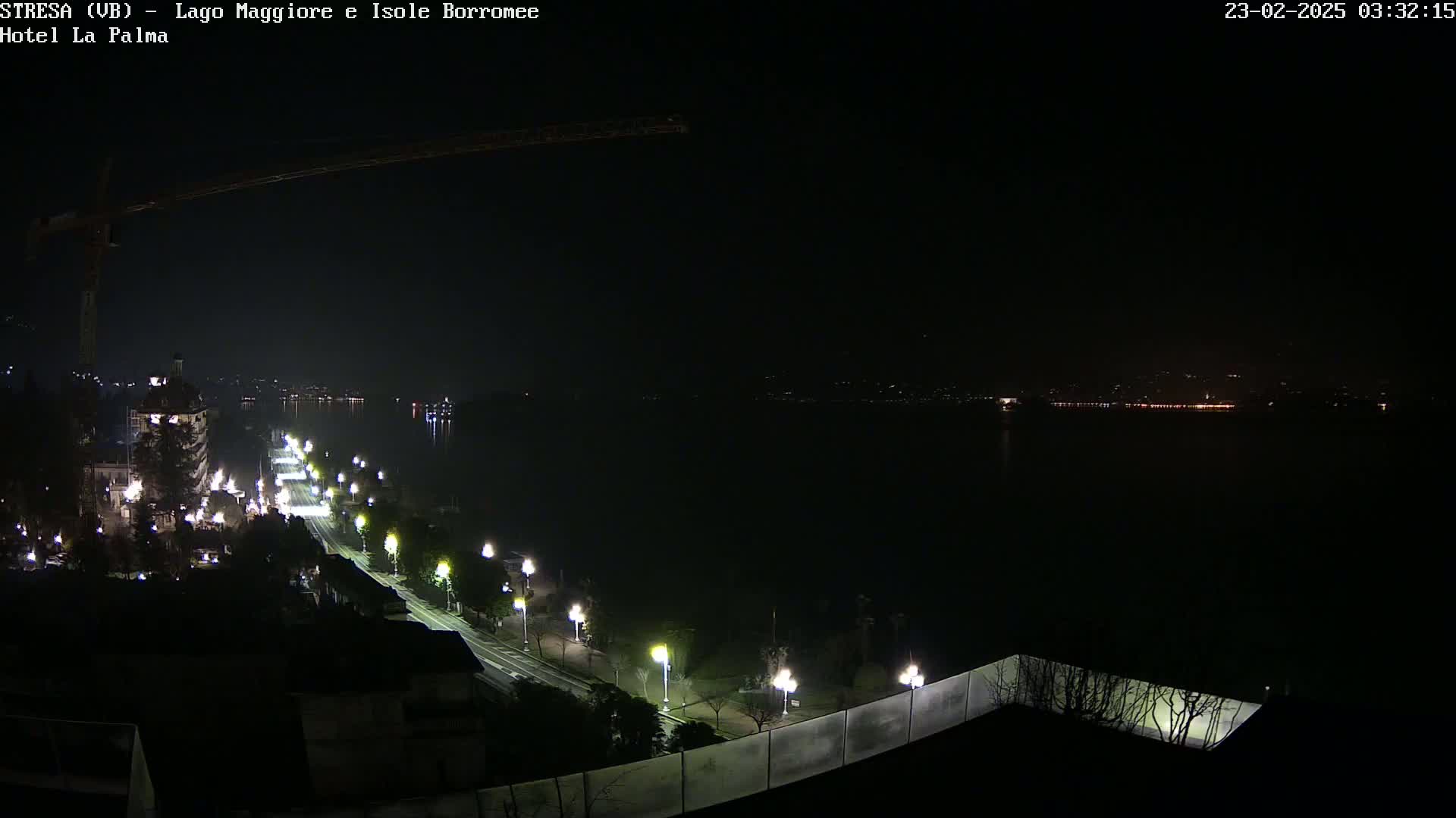 A nighttime view of a city street lit by lampposts along a body of water, with a construction crane visible in the upper left corner and a distant cityscape faintly illuminated across the water.