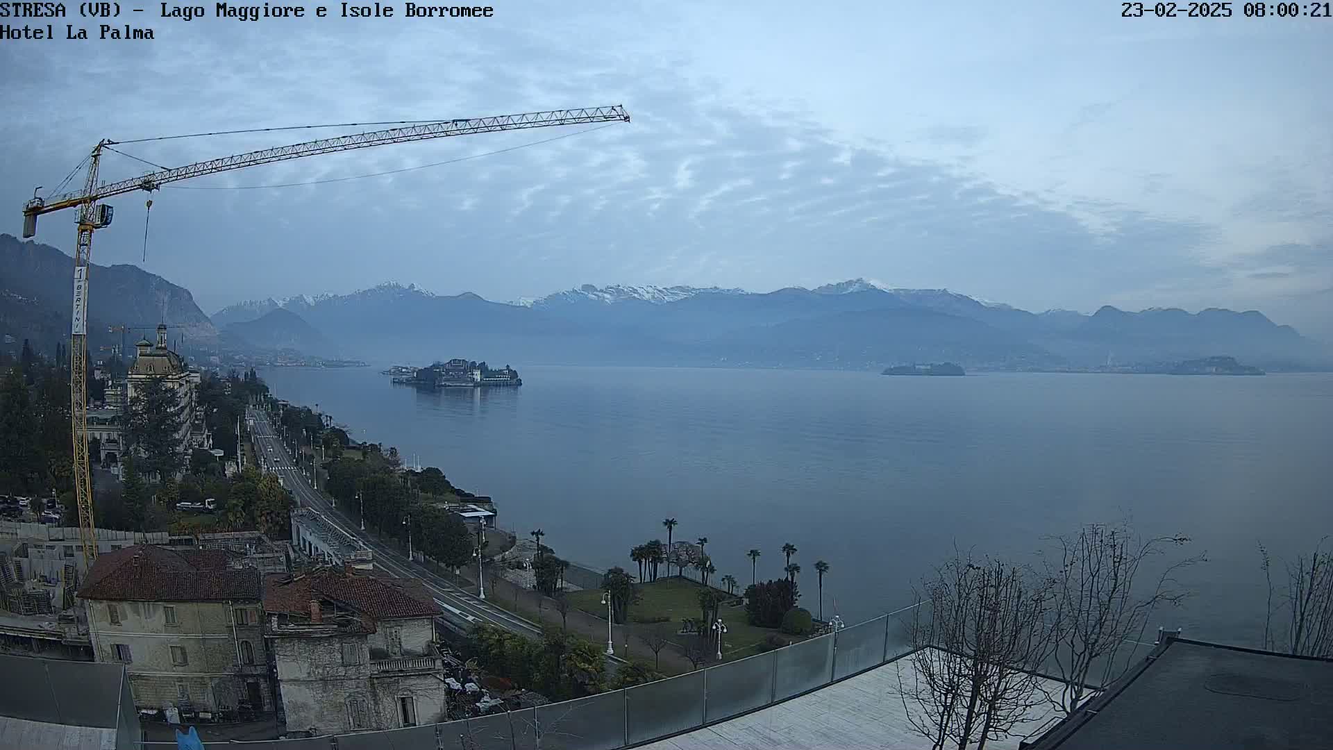 A large construction crane overlooks a calm lake with snow-capped mountains in the distance under a mostly cloudy sky.