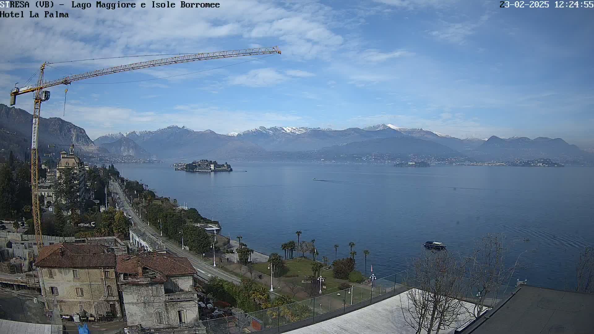 A large lake is seen with snow-capped mountains in the background under a mostly clear sky, a construction crane is visible in the foreground, along with buildings and a shoreline road.