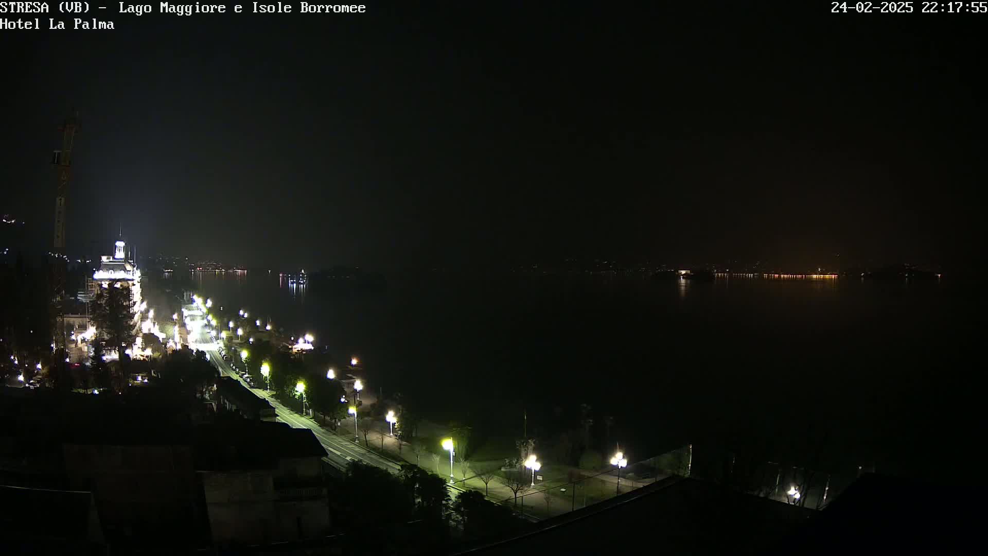 A nighttime view of a city street lined with lights along a dark body of water, with faint lights visible on the opposite shore.
