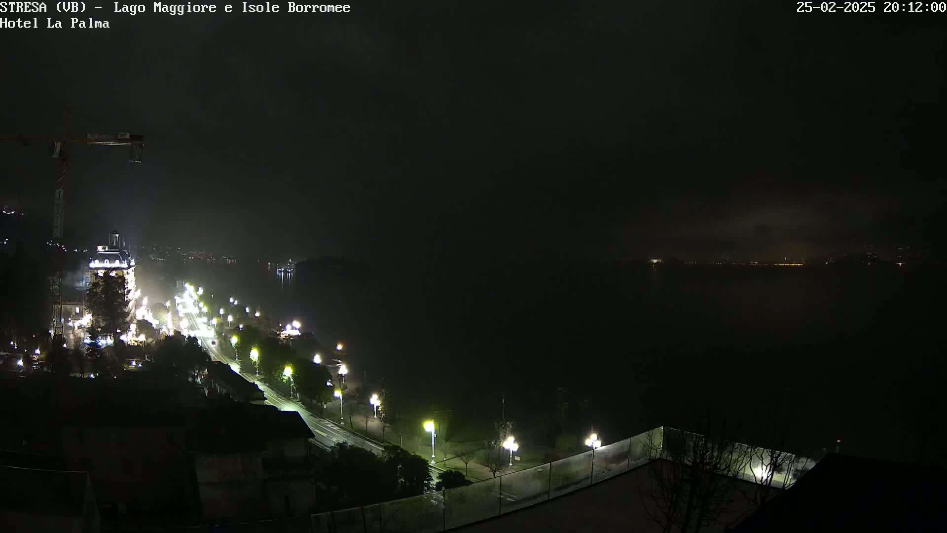 A nighttime, foggy view of a city street lined with lights leading to a dark body of water with faintly visible lights on the opposite shore, a construction crane is visible near a building to the left.