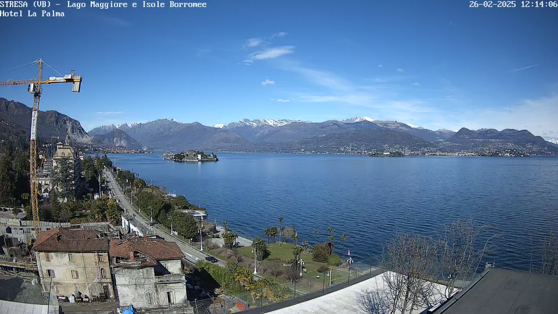A clear, sunny day overlooking a lake with snow-capped mountains in the background, a small island visible in the middle distance, and a construction crane near a shoreline with buildings.