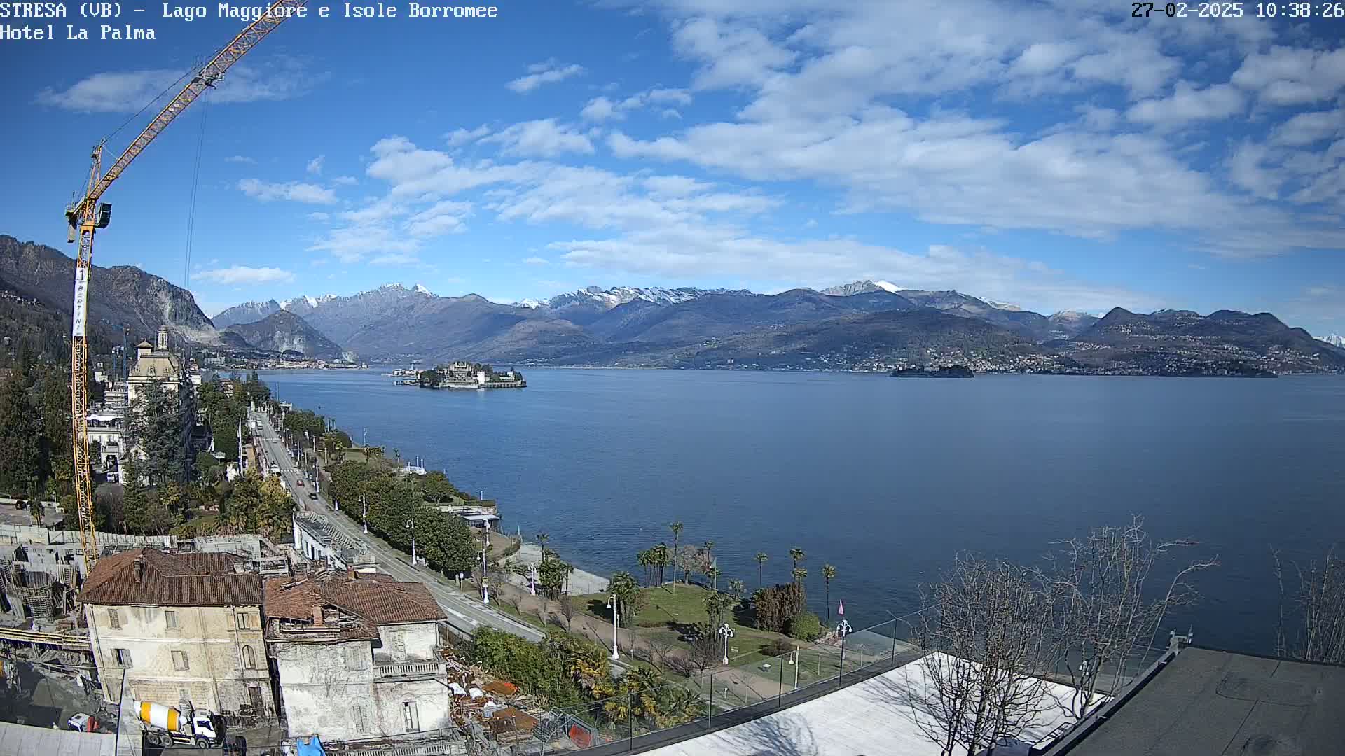 A partly sunny day overlooks a large lake with snow-capped mountains in the distance, a construction crane, and a shoreline with buildings and palm trees.